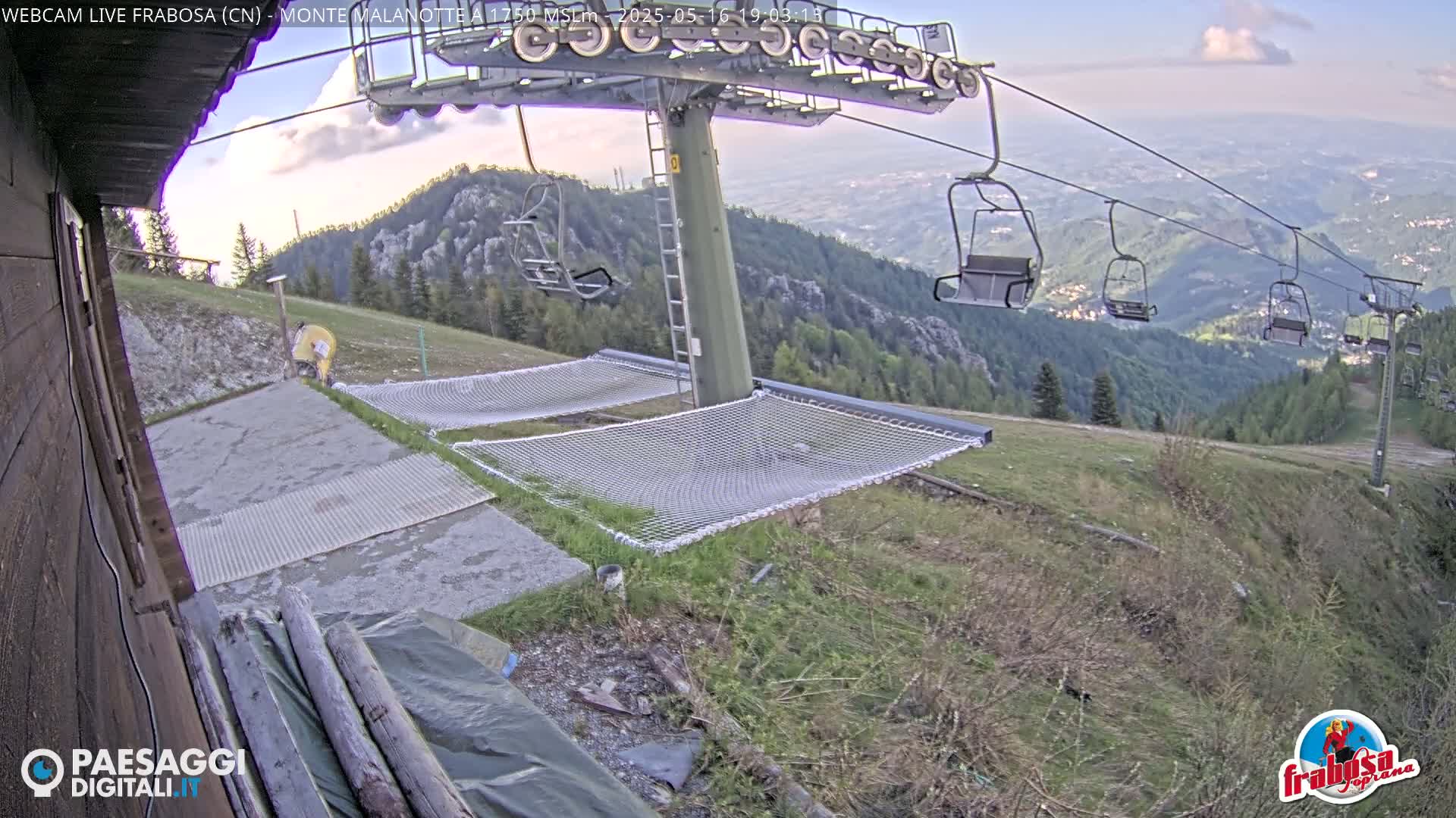 A mountaintop view shows a chairlift with empty chairs, two empty net hammocks, and a grassy area, under partly cloudy skies.