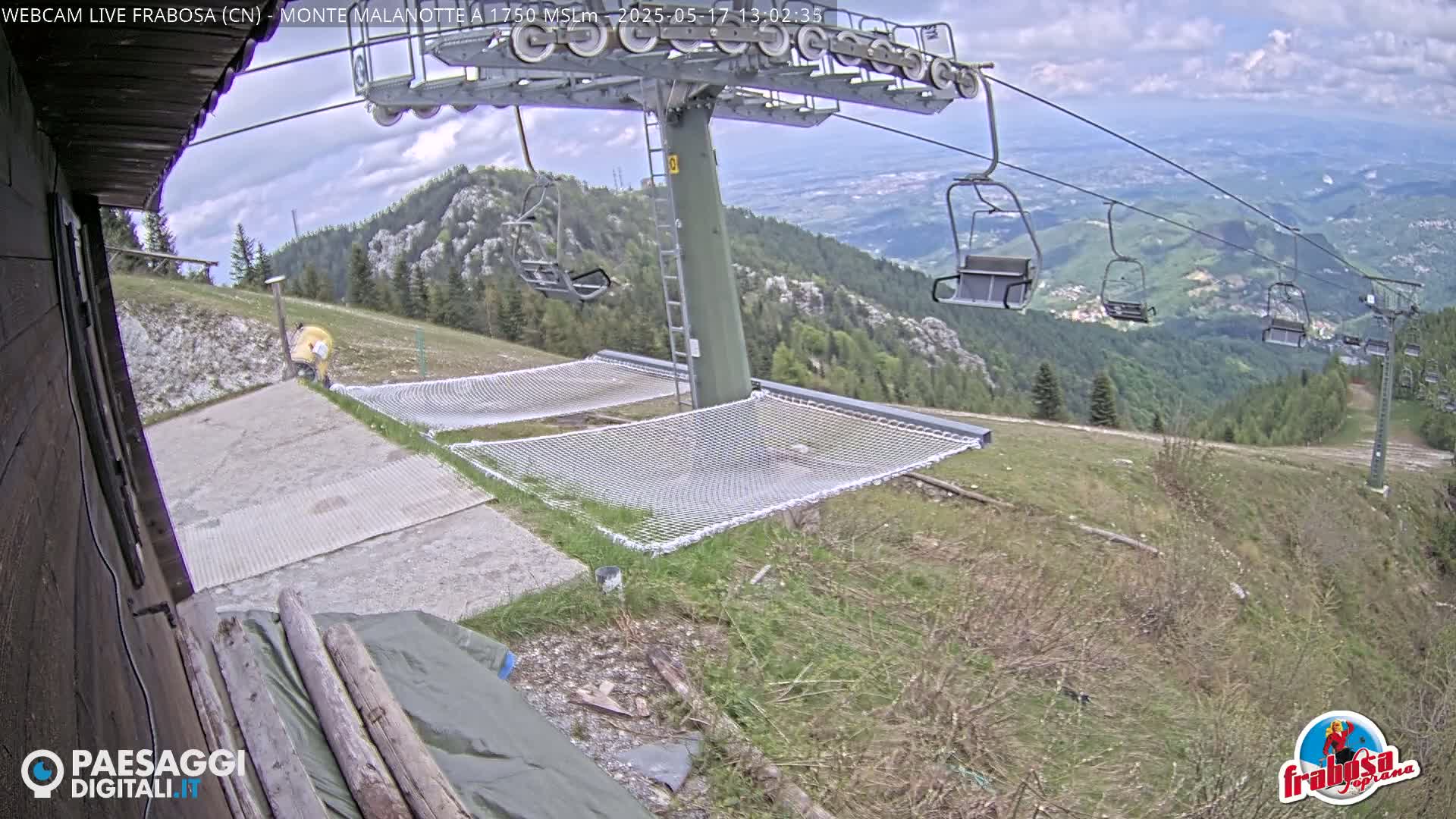 A mountaintop view shows a chairlift, several empty chairlift seats, and two large nets laid out on the ground under partly cloudy skies.