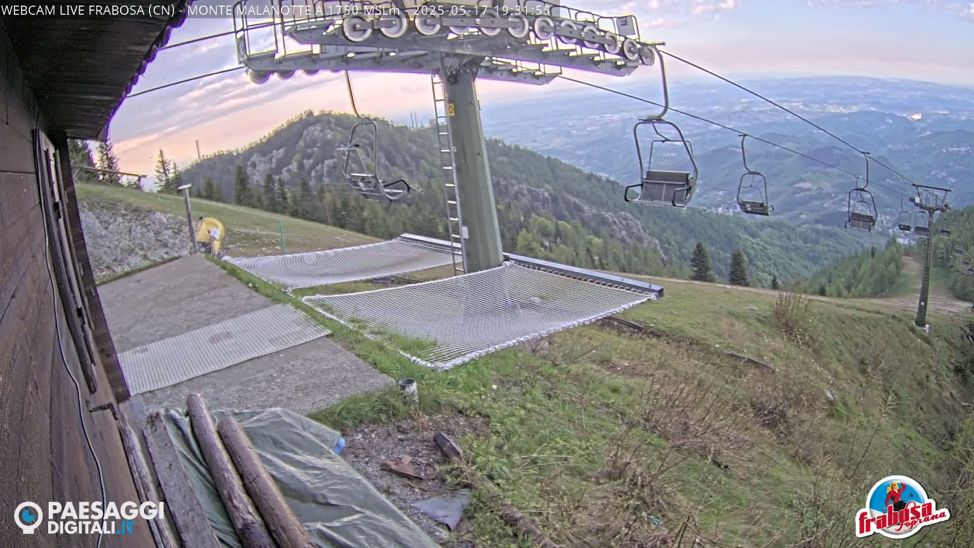 A mountaintop view shows several empty chairlifts against a partly cloudy evening sky, with grassy slopes and safety nets near a mountain lodge.
