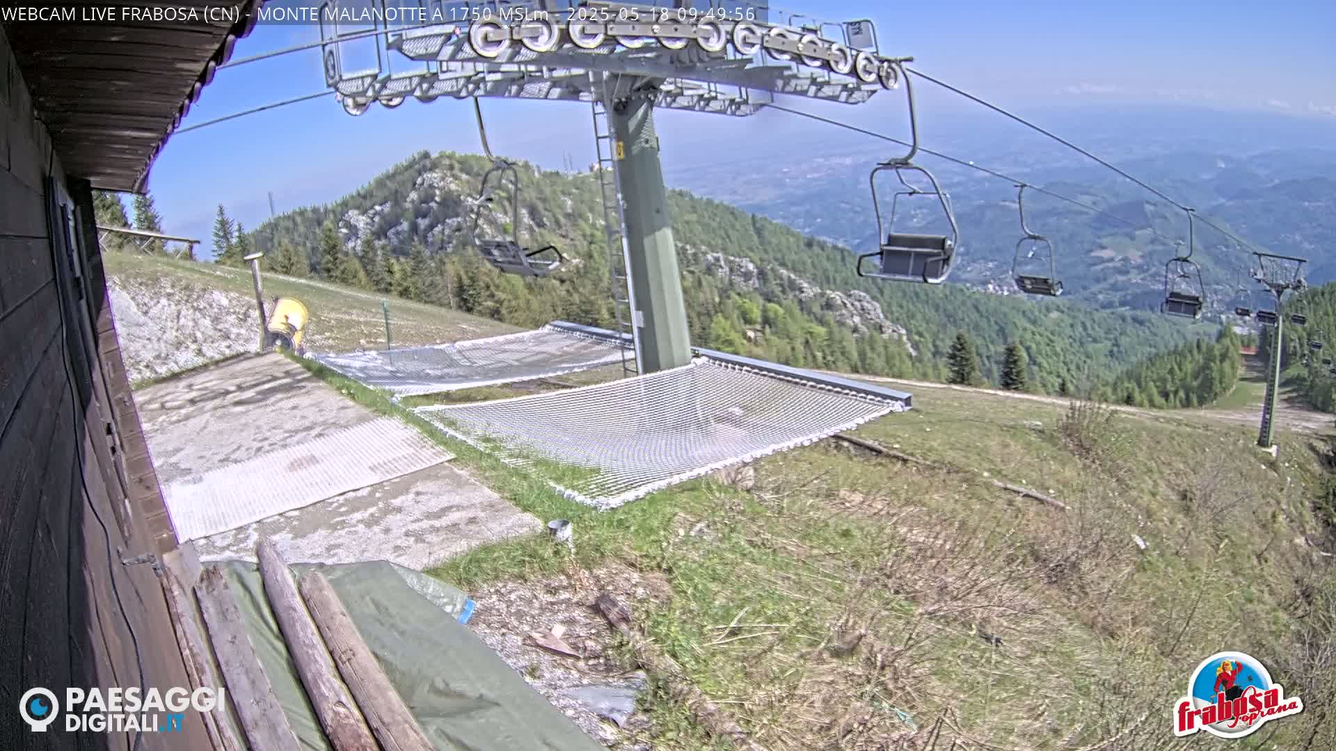 A sunny mountaintop scene shows a chairlift, two empty safety nets, and a grassy area overlooking a valley.