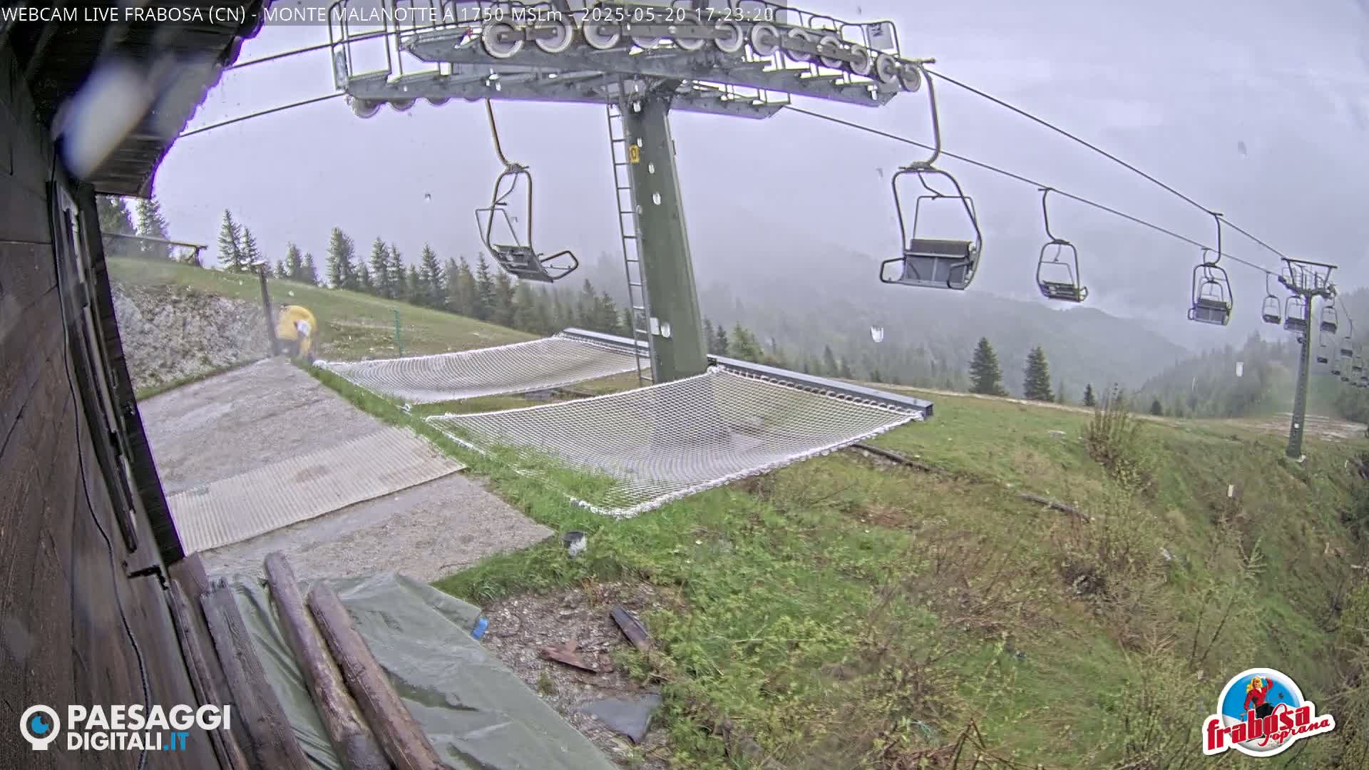A ski lift with empty chairs is seen in the rain, with safety nets below, and a person in yellow is walking near the bottom of the hill.