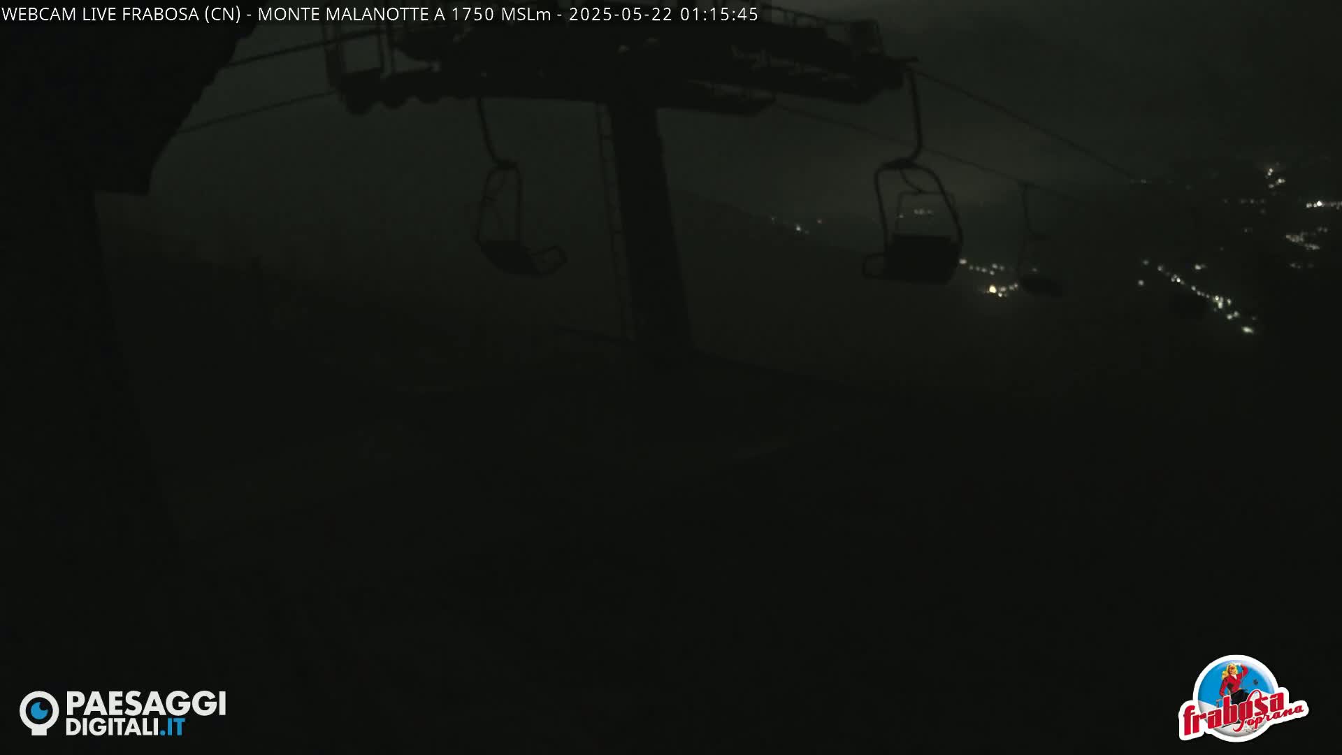 At night, empty chairlifts hang against a dark, blurry background of distant lights.