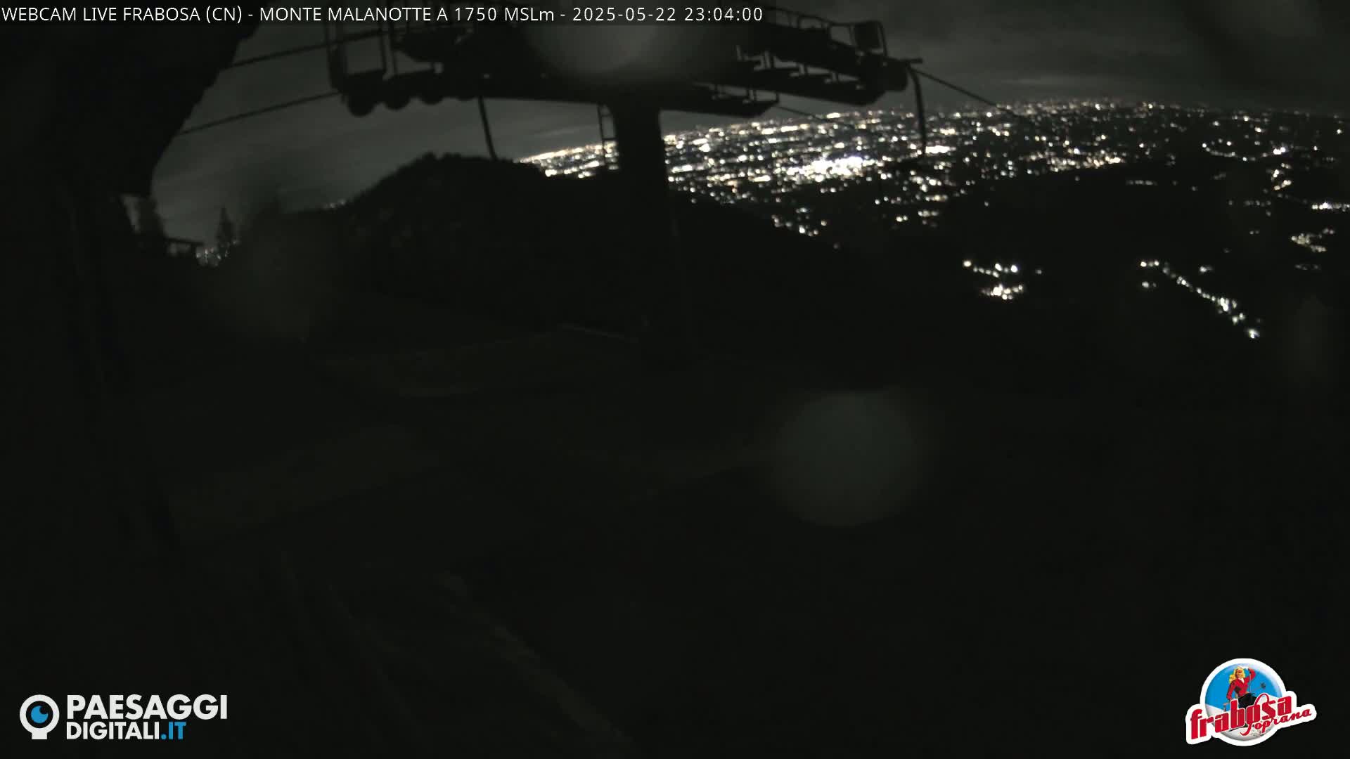 A nighttime view from a mountaintop shows a distant city illuminated below, partially obscured by darkness and a chairlift in the foreground.