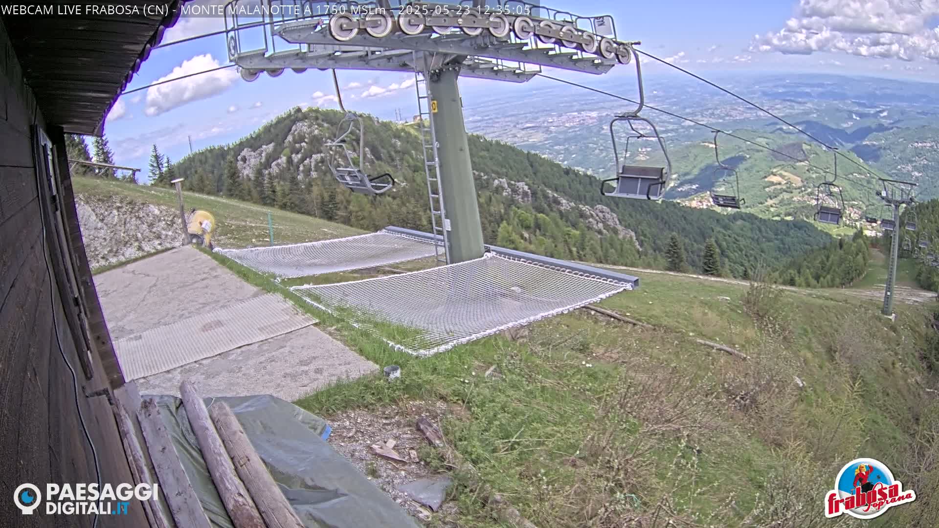 A chairlift on a mountaintop with partly cloudy skies overlooks a valley, and several safety nets are on the ground below.