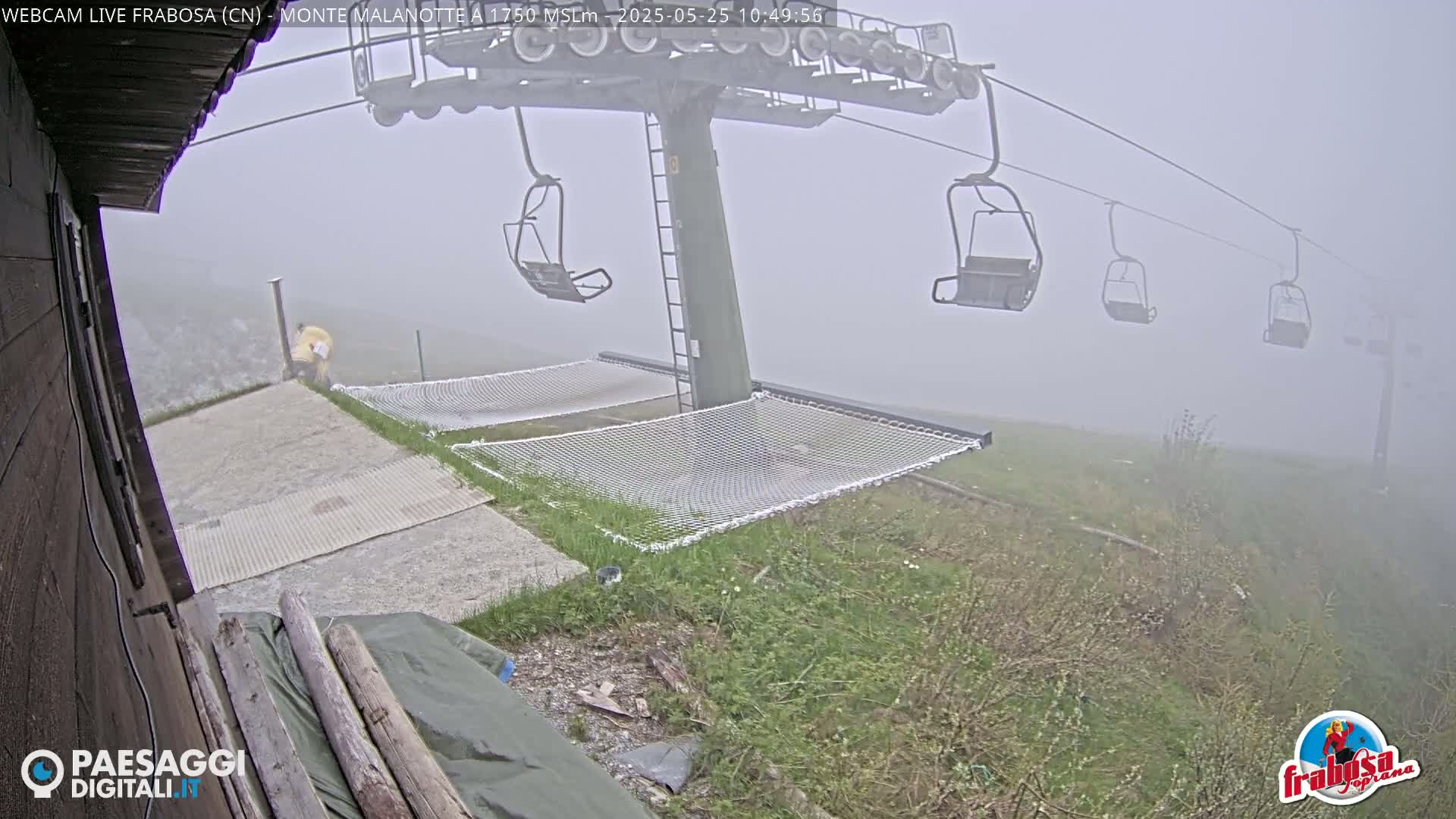 A chairlift with empty chairs stands in thick fog above grassy ground with safety nets, near a building.