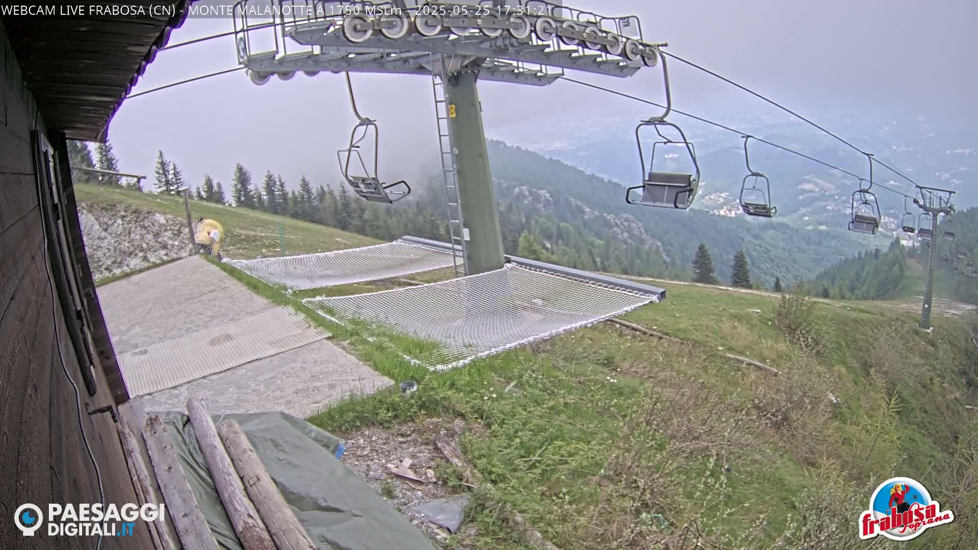 A chairlift ascends a mountainside on an overcast day, with empty chairs visible and several net hammocks near a paved area.
