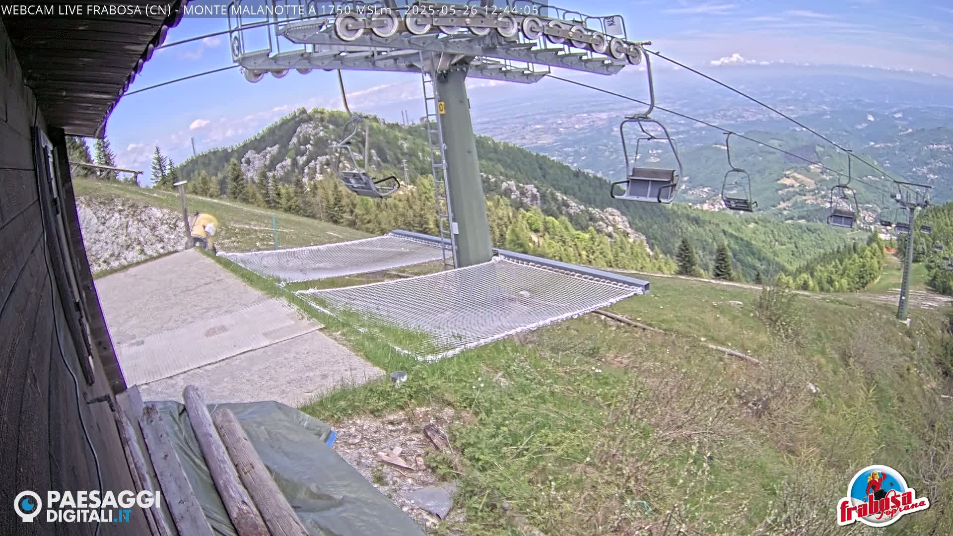 A mountaintop ski lift with empty chairs overlooks a valley on a sunny day, with grassy areas and netting near the lift's base.