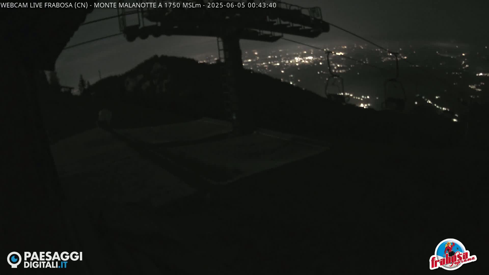 A nighttime view from a ski lift shows a dark landscape with city lights in the distance under a clear sky.