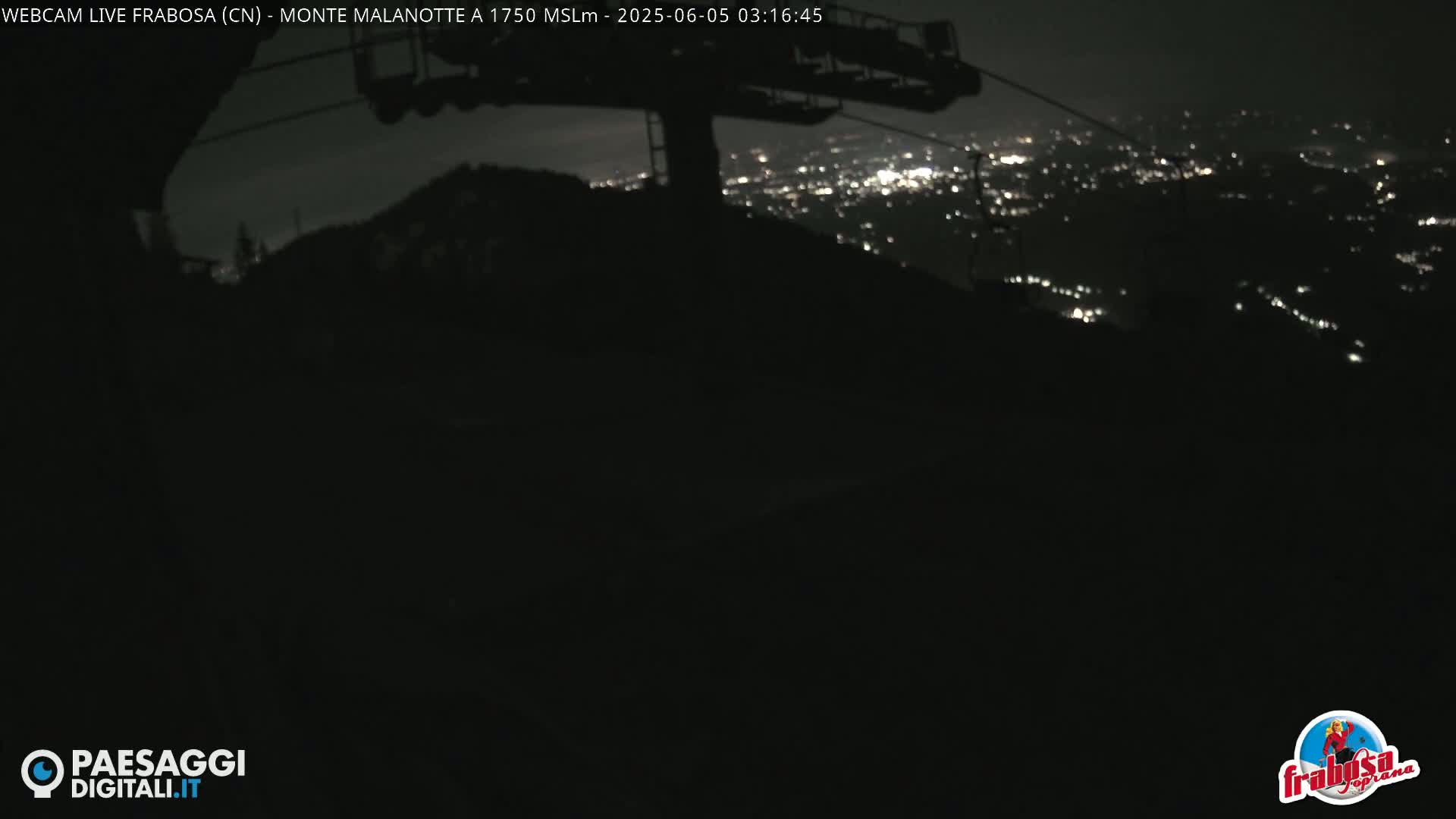 A nighttime view from a mountaintop shows a chairlift and a distant city glowing with lights.
