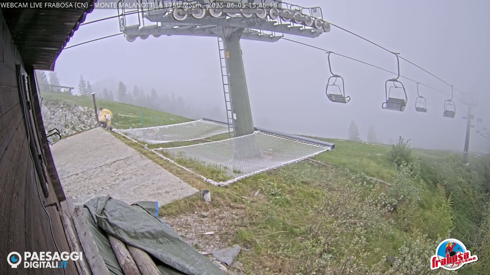 A foggy mountaintop scene shows an empty chairlift, several safety nets, and a paved area next to a wooden structure.
