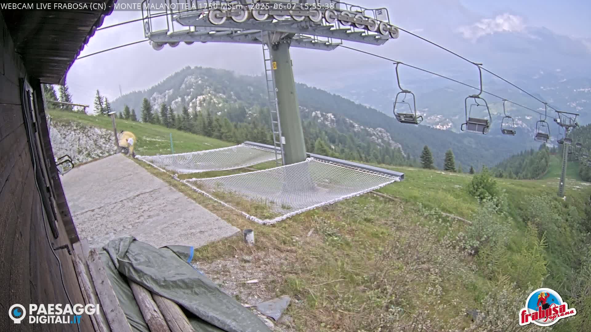 A chairlift ascends a mountainside on an overcast day, with several empty chairlift seats visible, and two empty mesh hammocks situated on a grassy area near a concrete pad.