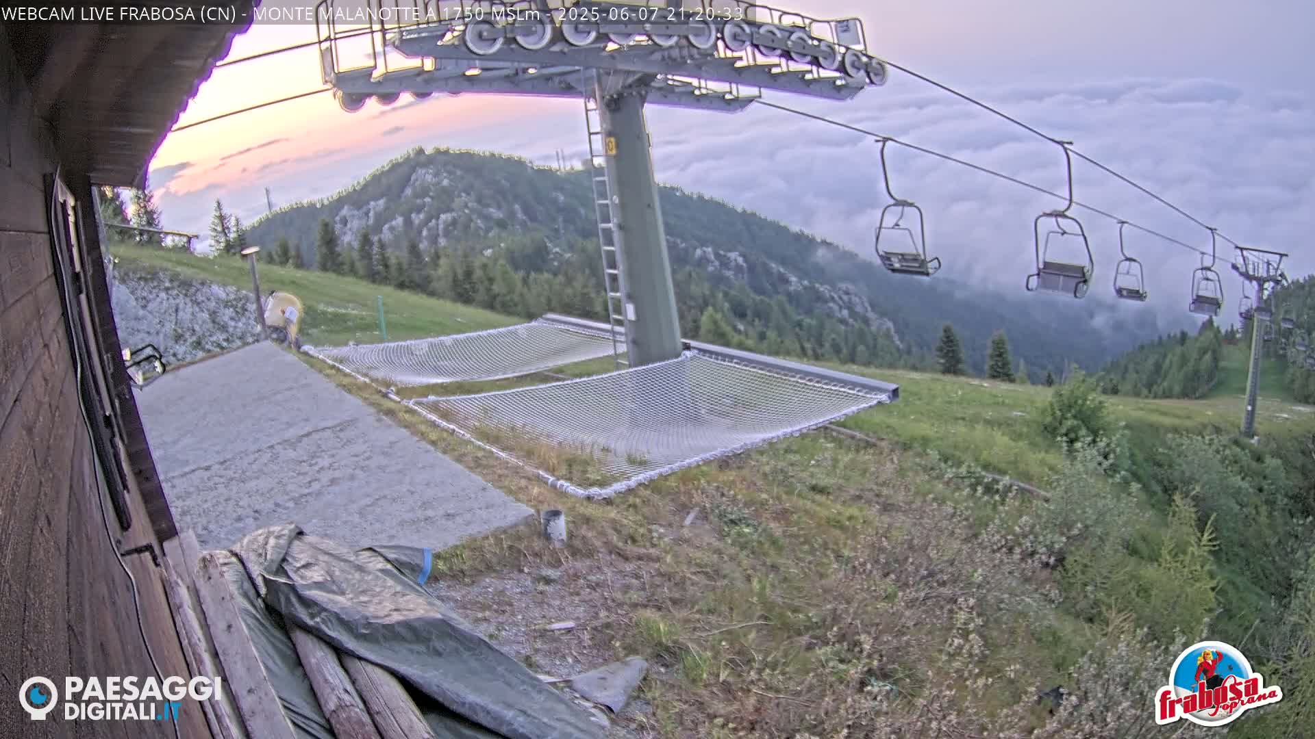 A chairlift ascends a mountain shrouded in low clouds at dusk, with two net hammocks resting on the ground nearby.