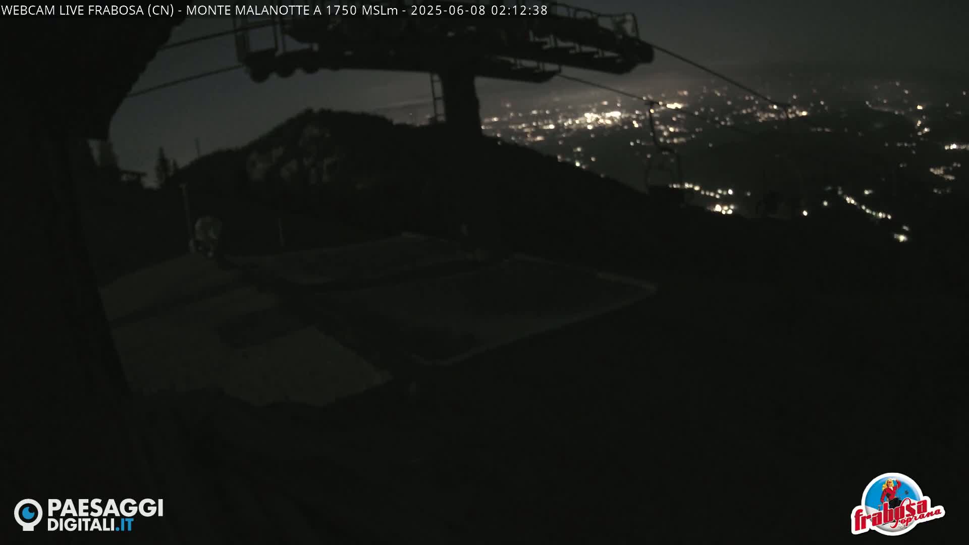 A nighttime view from a mountaintop shows a chairlift and a distant city illuminated below.