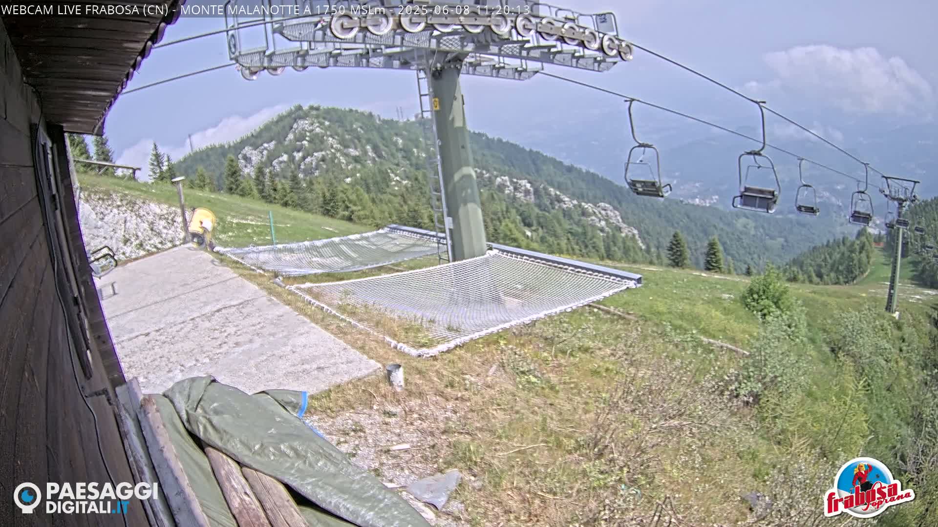 A mountaintop view shows a chairlift, several empty chairs, and two large nets resting on the ground, all under a mostly clear, sunny sky.