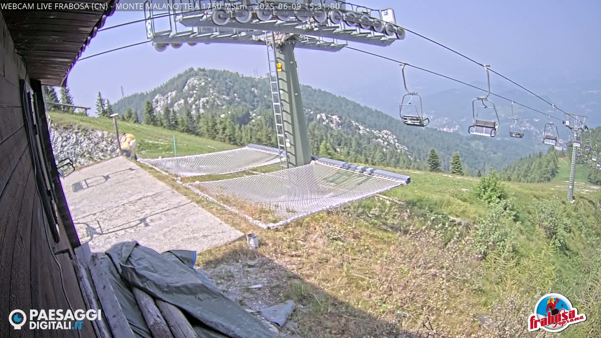A chairlift ascends a mountainside on a clear, sunny day, with empty chairs visible, and two empty net hammocks situated near a paved area at the base of the lift.