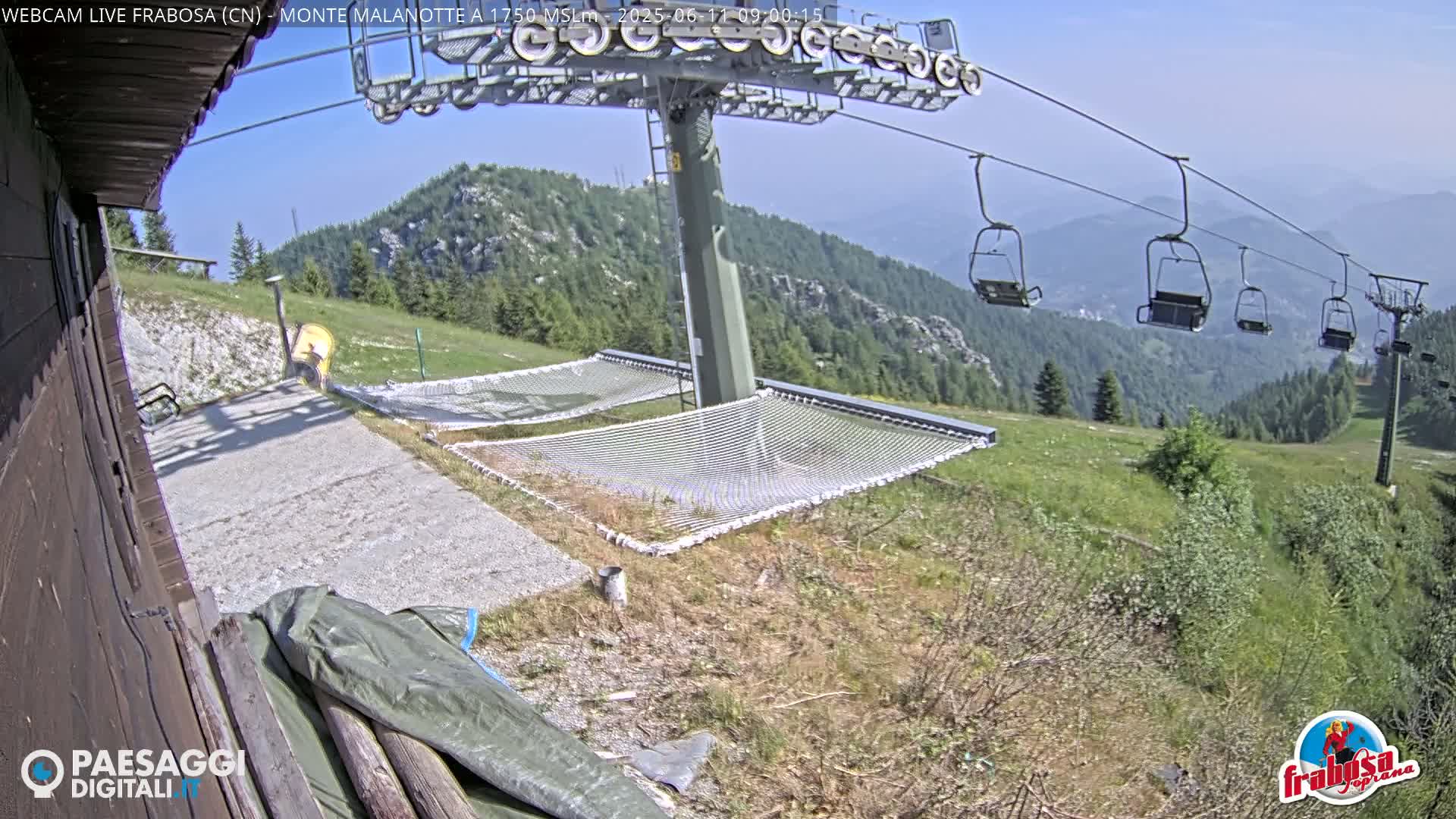 A mountaintop scene on a clear, sunny day shows a chairlift, several empty chairlift seats, and two empty mesh hammocks near a building.