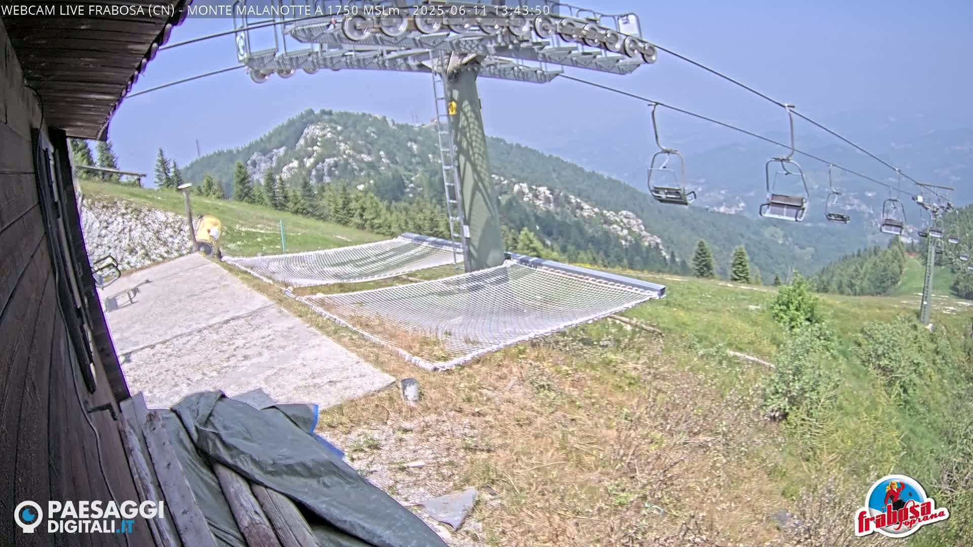 A mountaintop view shows a chairlift with empty seats, two empty net hammocks, and a grassy area under a clear, sunny sky.