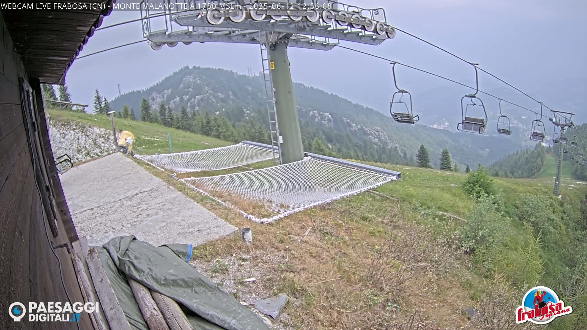 A chairlift ascends a mountain on an overcast day, passing over a grassy area with empty hammocks near a paved landing.