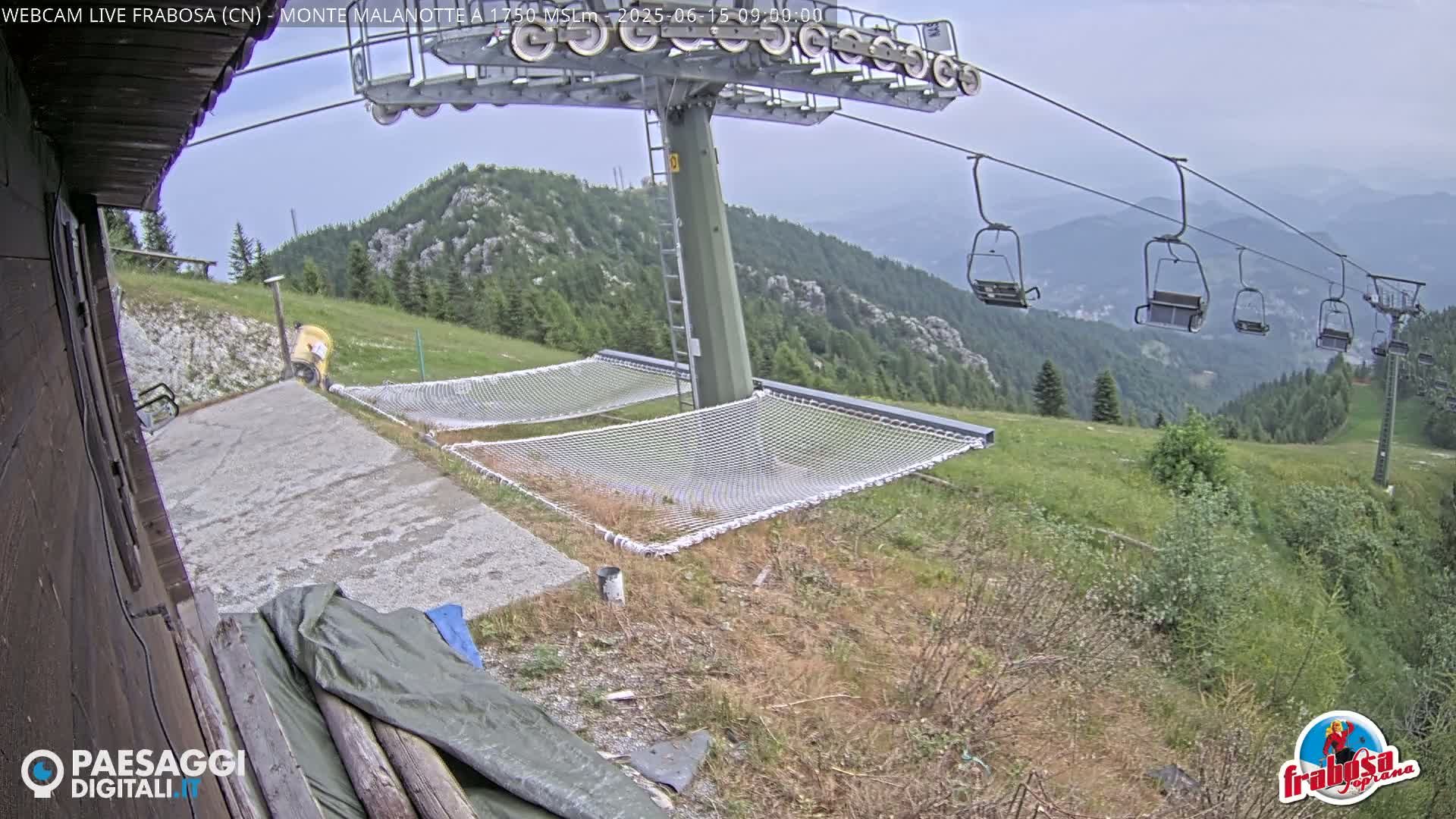 A mountaintop view on an overcast day shows a chairlift with empty chairs, several empty net hammocks, and a person in yellow near a building.