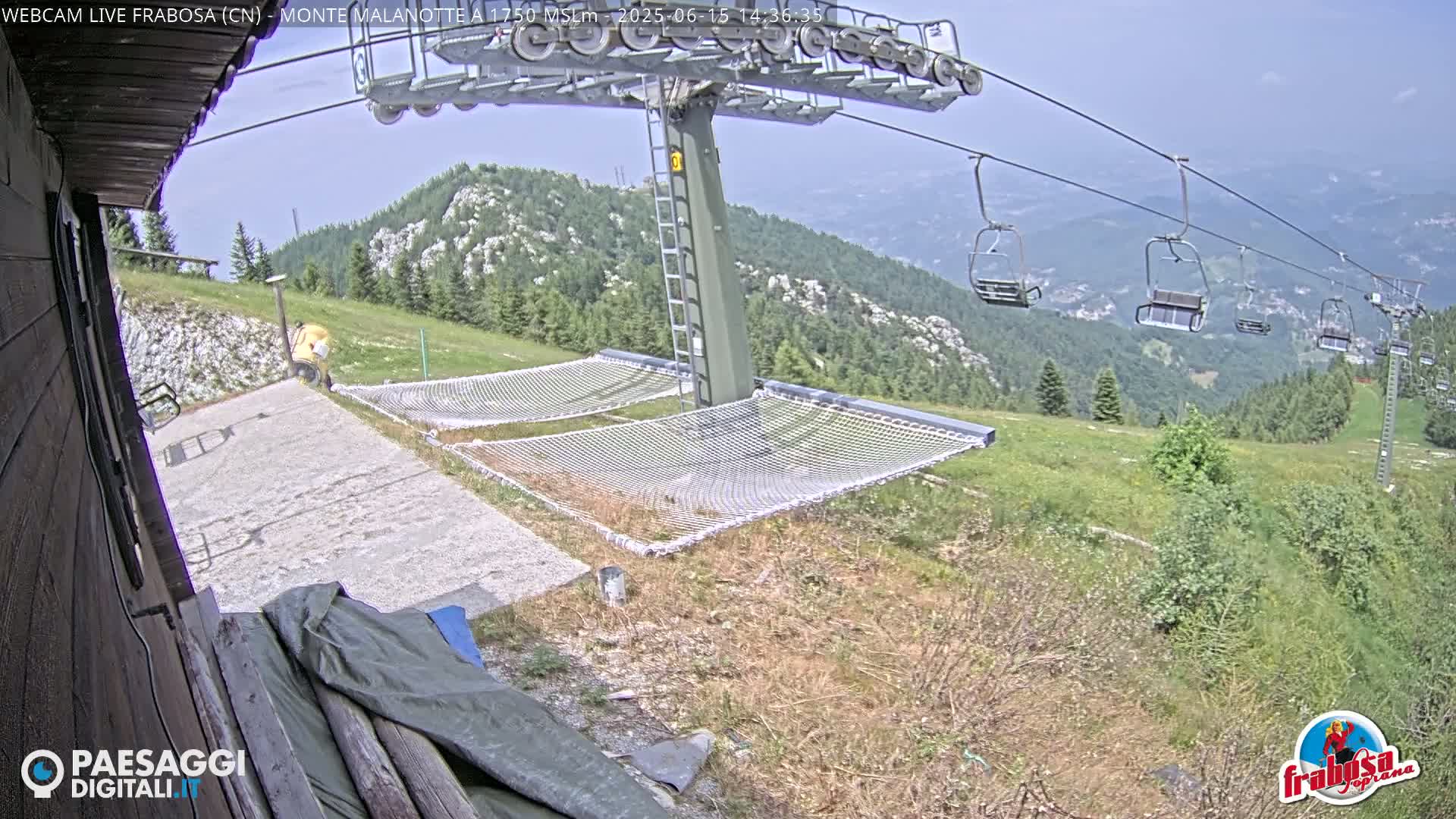 A mountaintop view shows a chairlift, empty net hammocks, and a person in the distance on a sunny day.