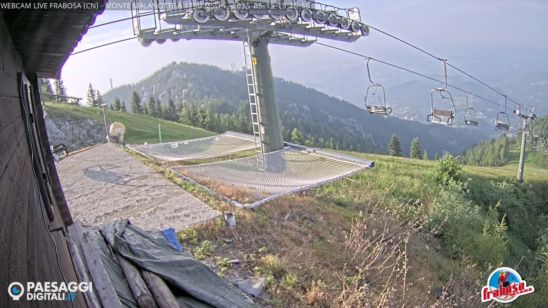A mountaintop view shows a chairlift, empty chairlift seats, and safety nets near a gravel area on a clear day.