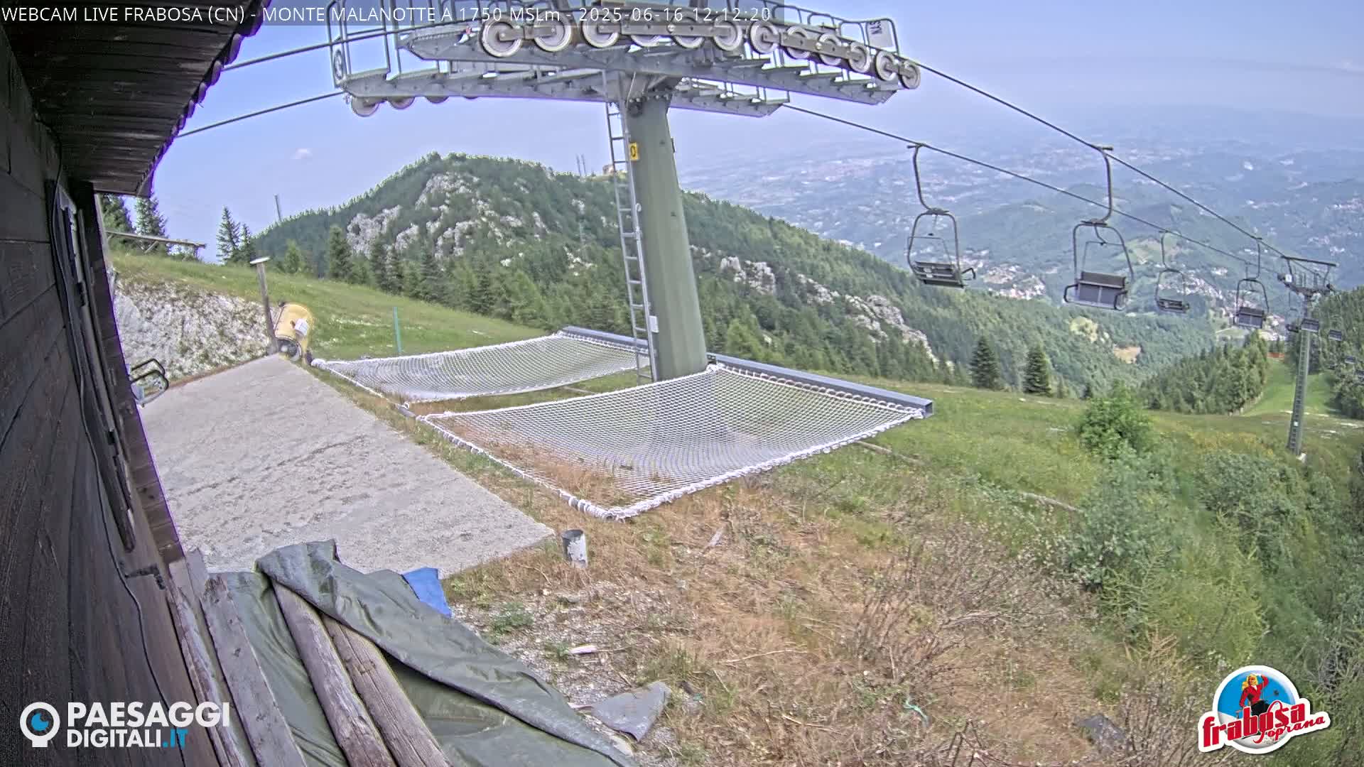 A mountaintop view shows a chairlift, two empty net hammocks, and a gravel area next to a wooden building, on a mostly clear day.