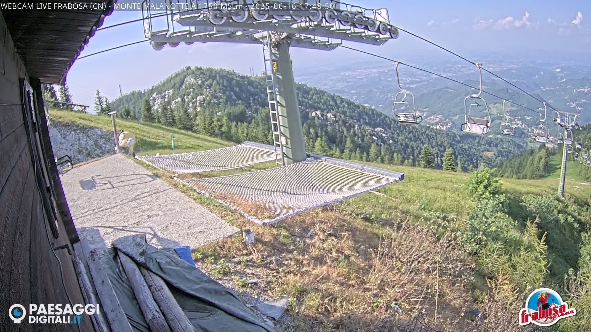 Two empty net hammocks sit near a chairlift station on a grassy mountainside under a mostly clear sky.