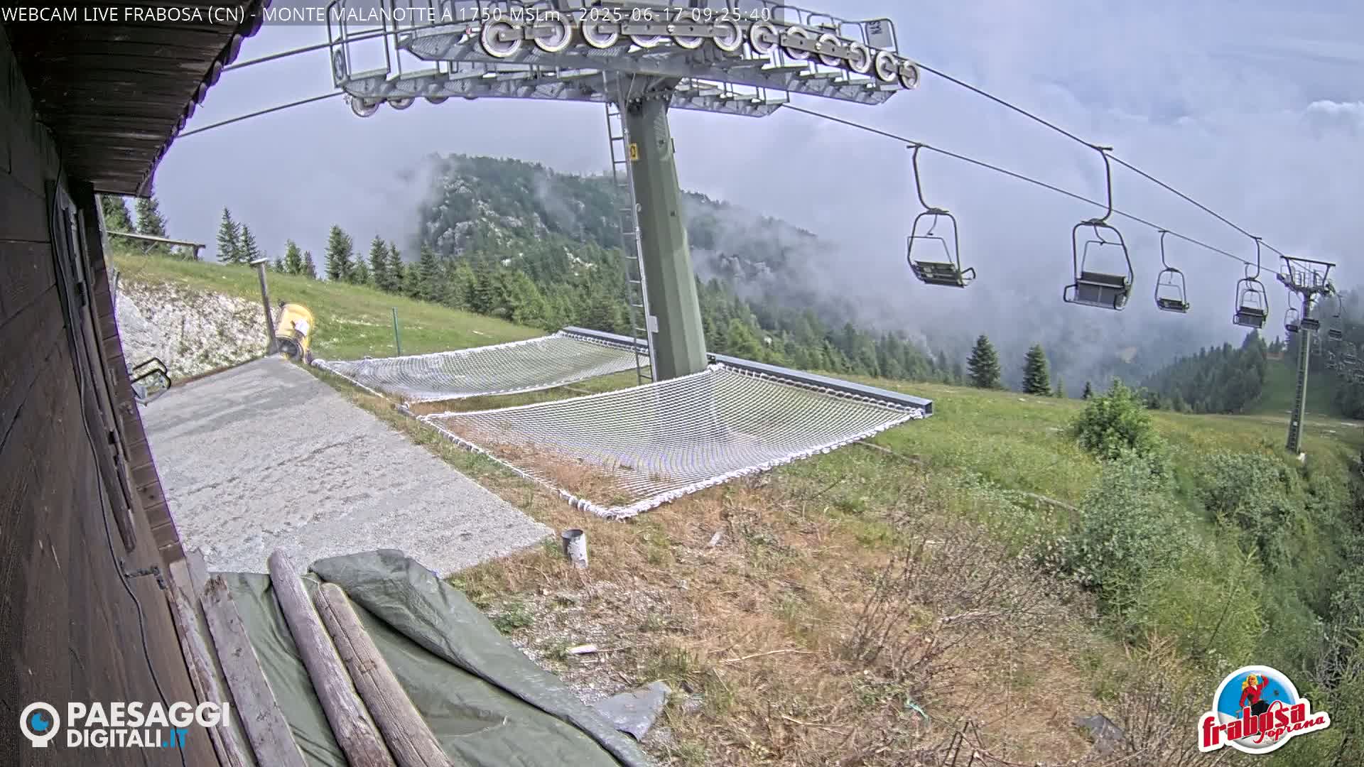 A mountaintop scene shows a chairlift with empty chairs, several empty net hammocks, a paved area, and a partly cloudy sky.