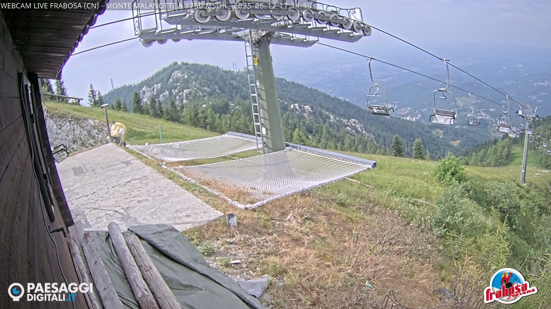 A mountaintop view shows a chairlift, two empty hammocks, and a person standing in the distance under a partly cloudy sky.