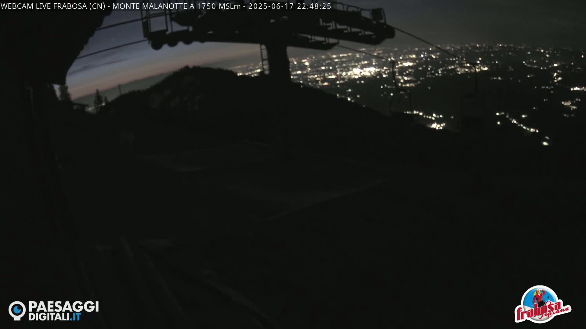 A nighttime view from a mountaintop shows a chairlift and a distant city glowing with lights under a dark sky.