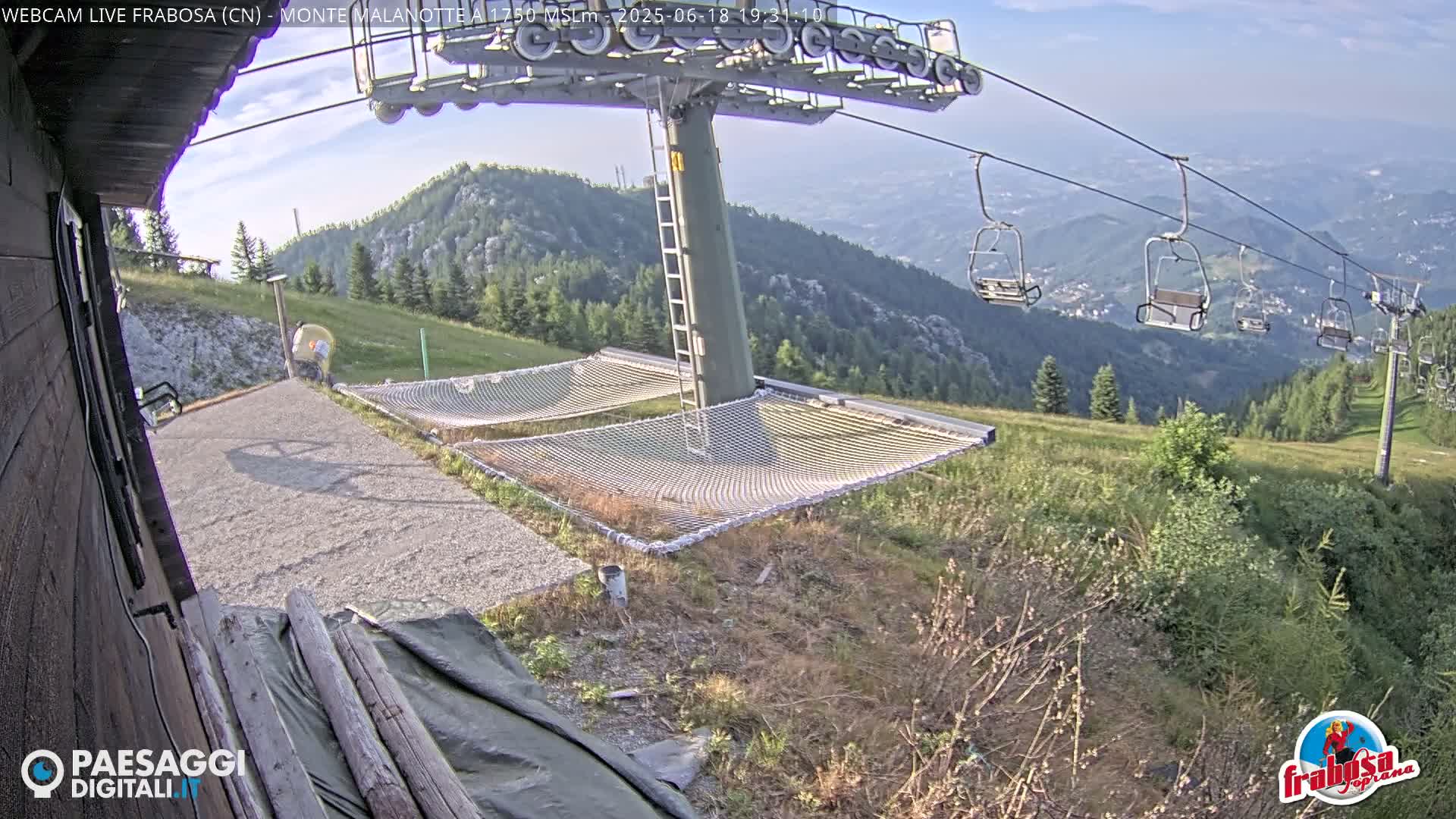 A mountaintop view shows a chairlift, several empty chairs hanging from cables, and two large netted hammocks on a gravel area next to a wooden building, all under a clear, sunny sky.