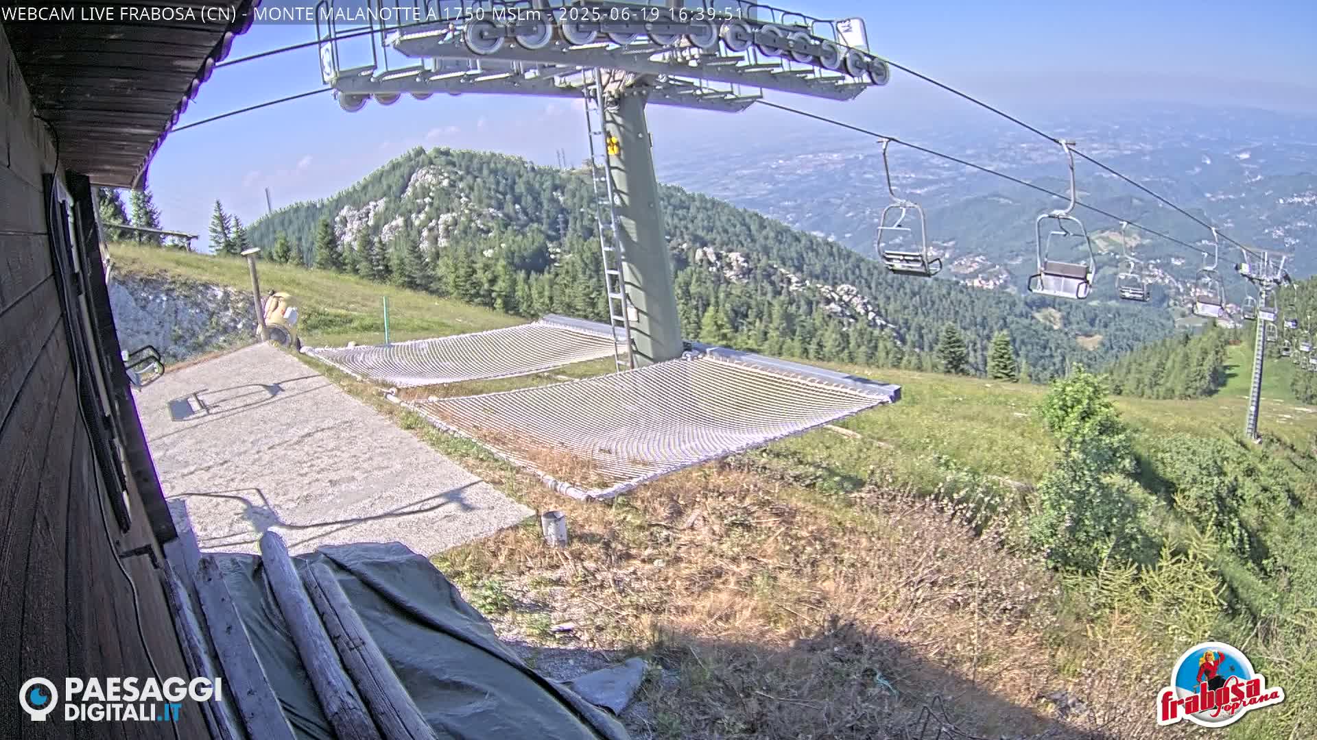 A sunny mountaintop view shows a chairlift, two empty net hammocks, and a person standing near a building, with a valley visible in the distance.