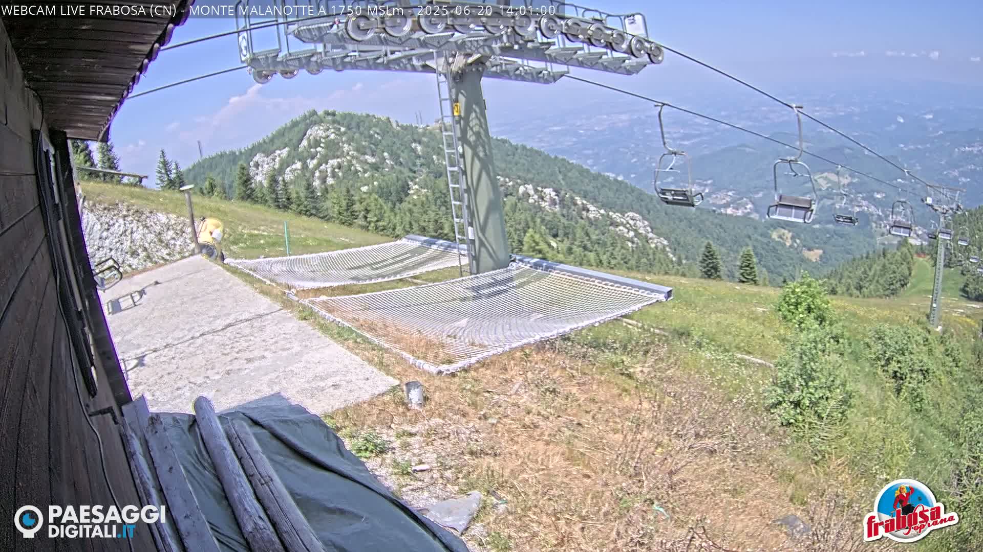 A mountaintop view on a sunny day shows a chairlift, two empty net hammocks, and a grassy hillside with distant mountain ranges and towns.