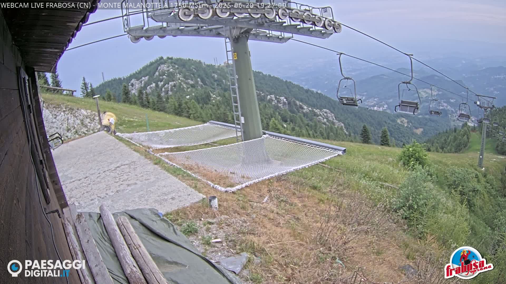 A mountaintop view shows a chairlift ascending a hill, with two empty net hammocks near a gravel area and a partly cloudy sky.