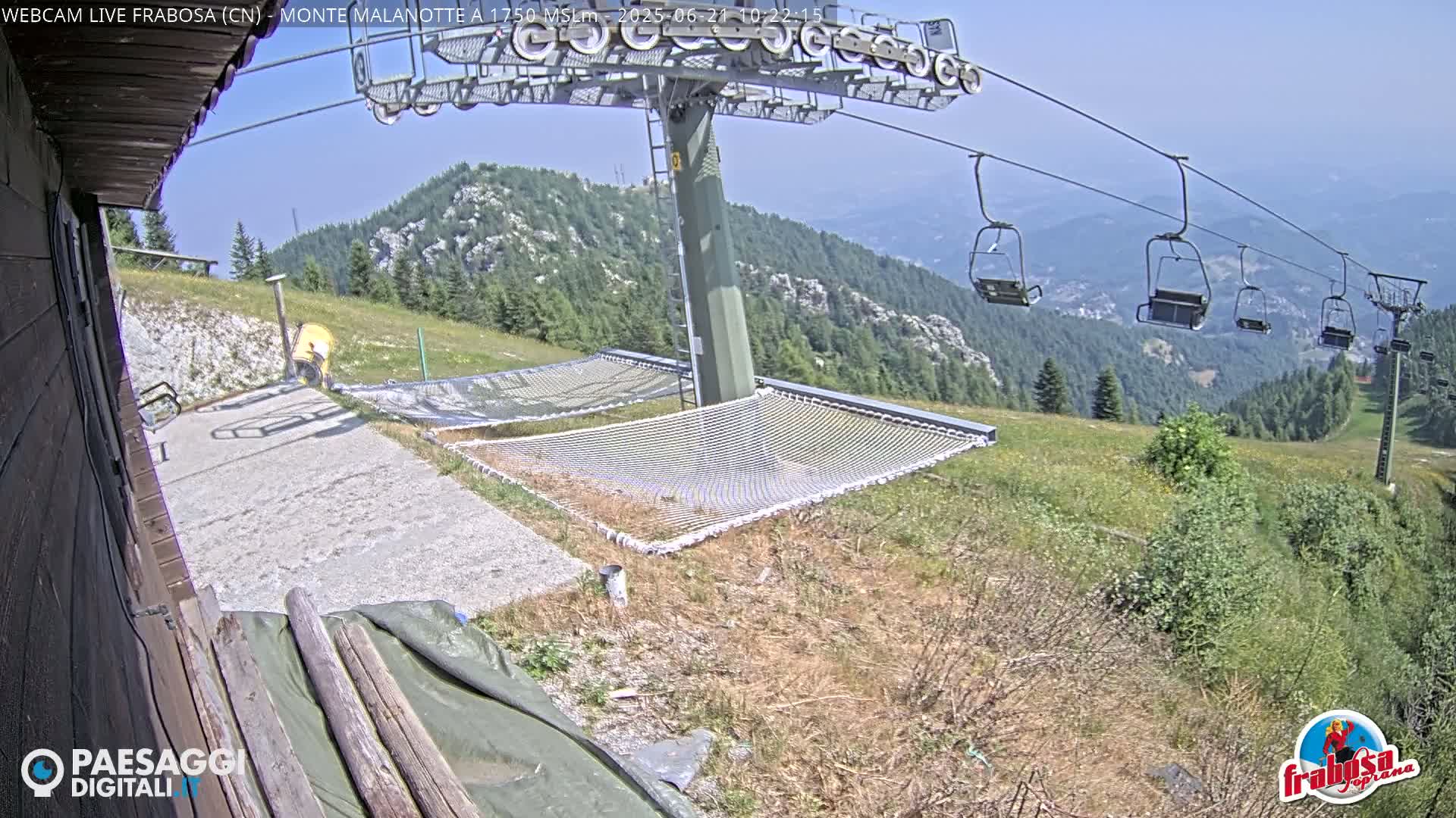 A mountaintop view shows a chairlift with empty seats, several large nets laid out on the ground, and a person standing near a building under a clear, sunny sky.