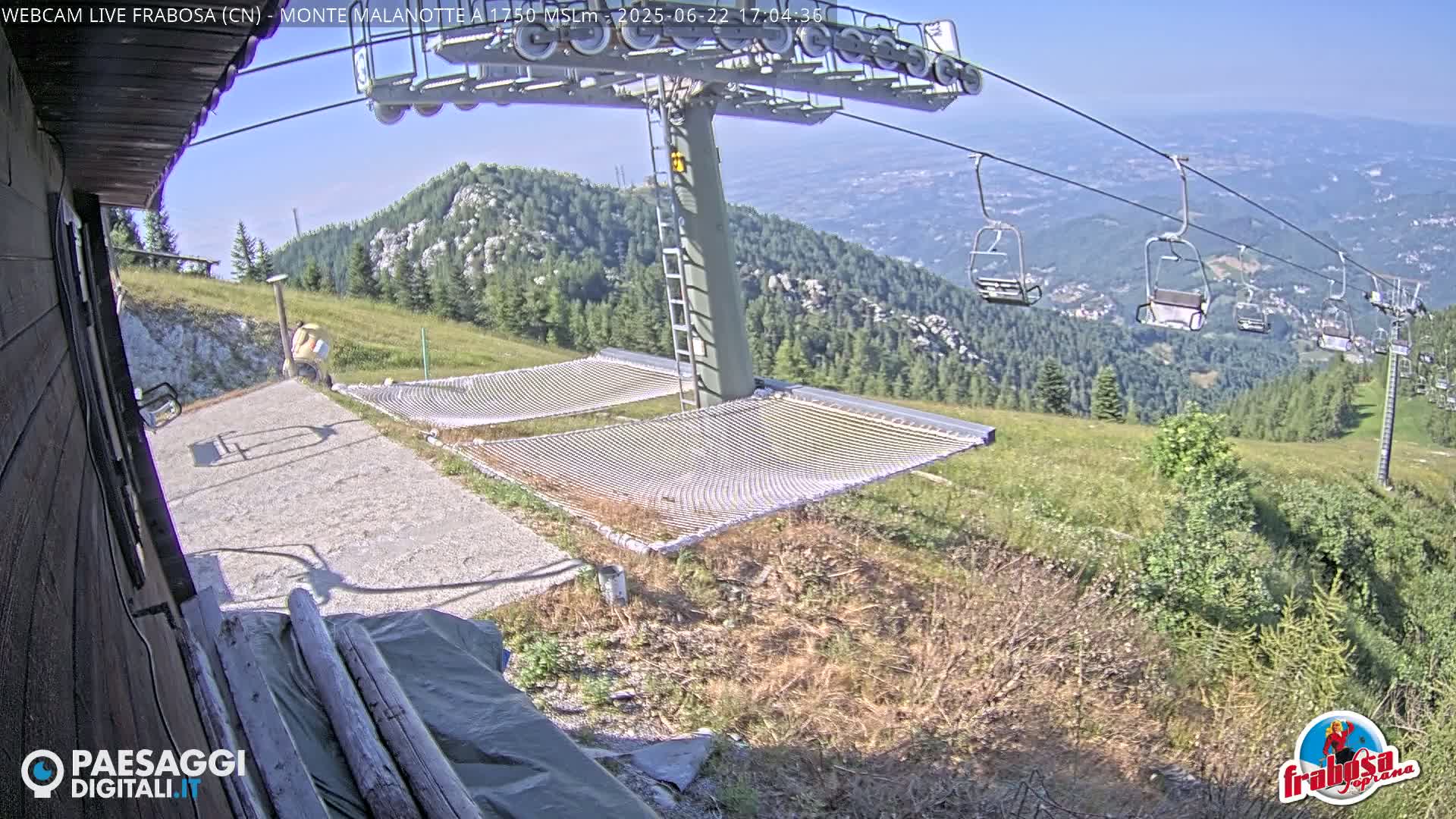 A mountaintop view shows a chairlift, two empty net hammocks, and a person standing near a building, all under a clear, sunny sky.