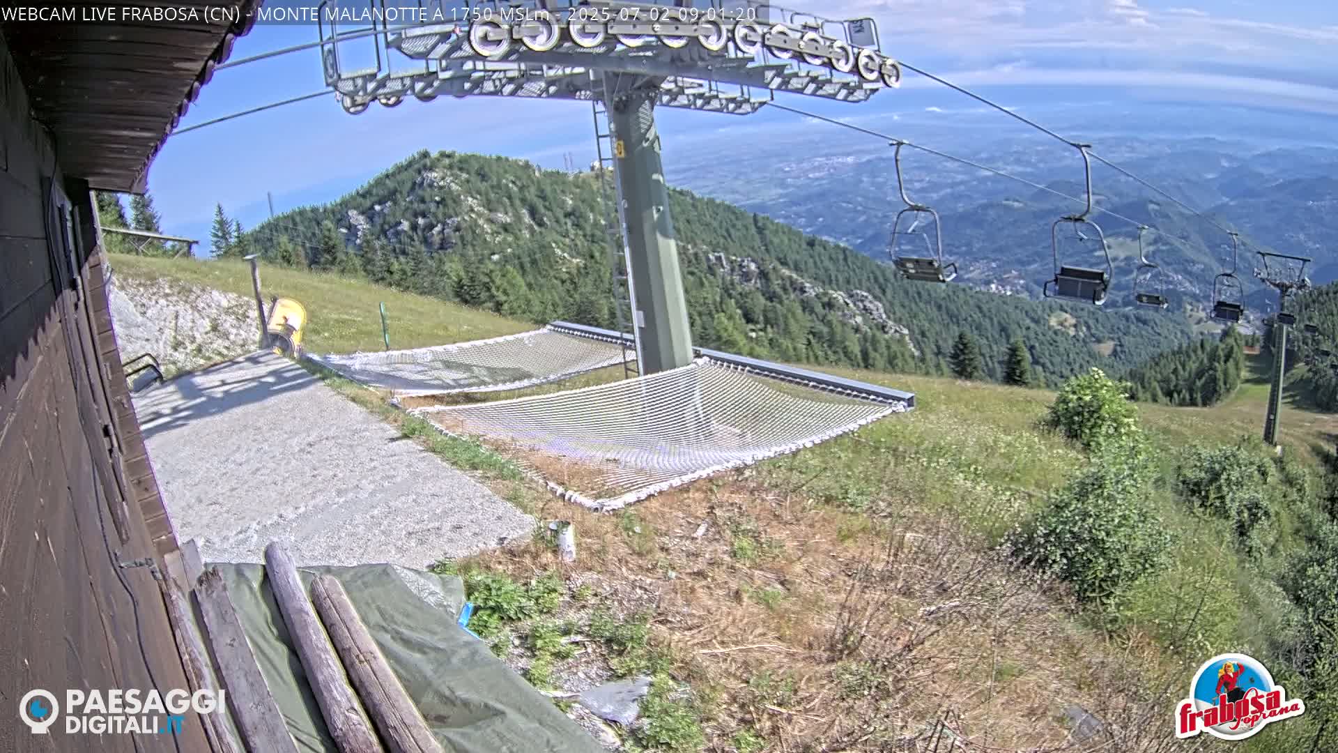 A mountaintop view on a sunny day shows two empty net hammocks near a chairlift, with a grassy hillside and distant valley visible.
