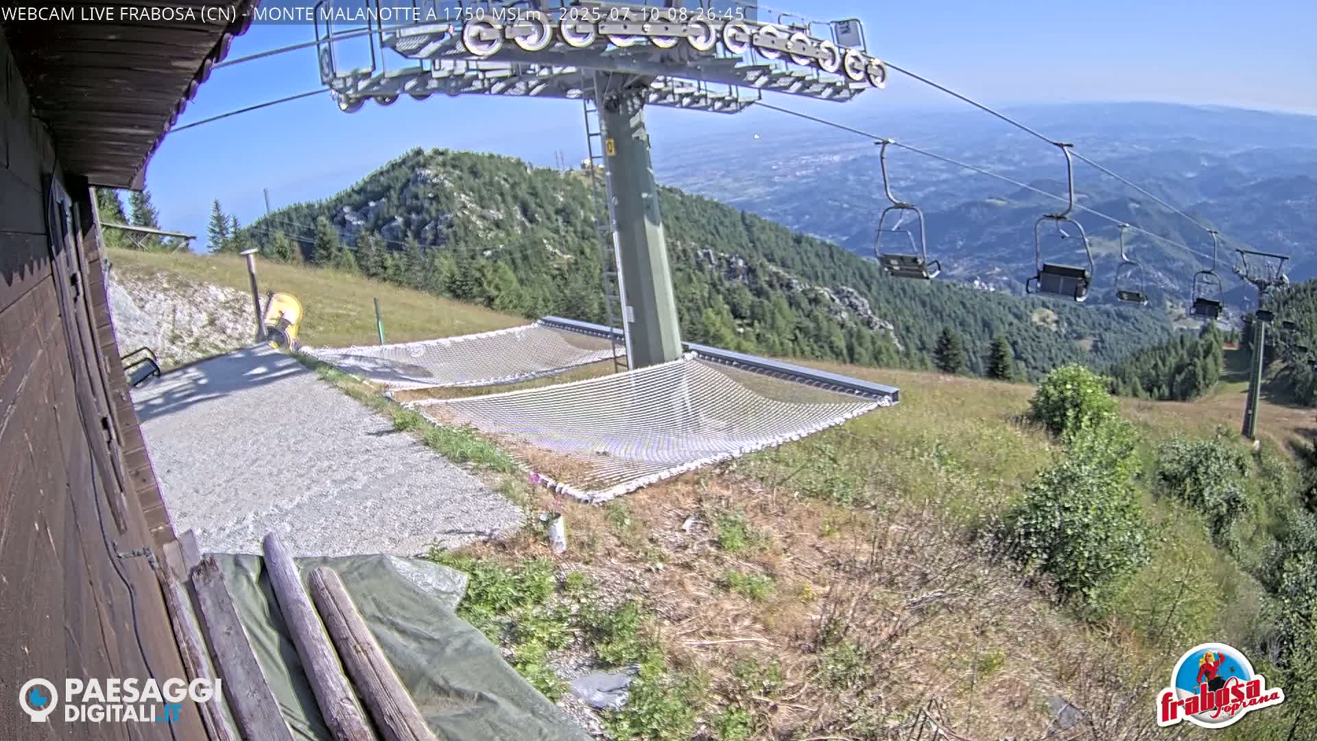 A mountaintop view on a sunny day shows a chairlift, two empty hammocks, and a gravel path next to a wooden building.