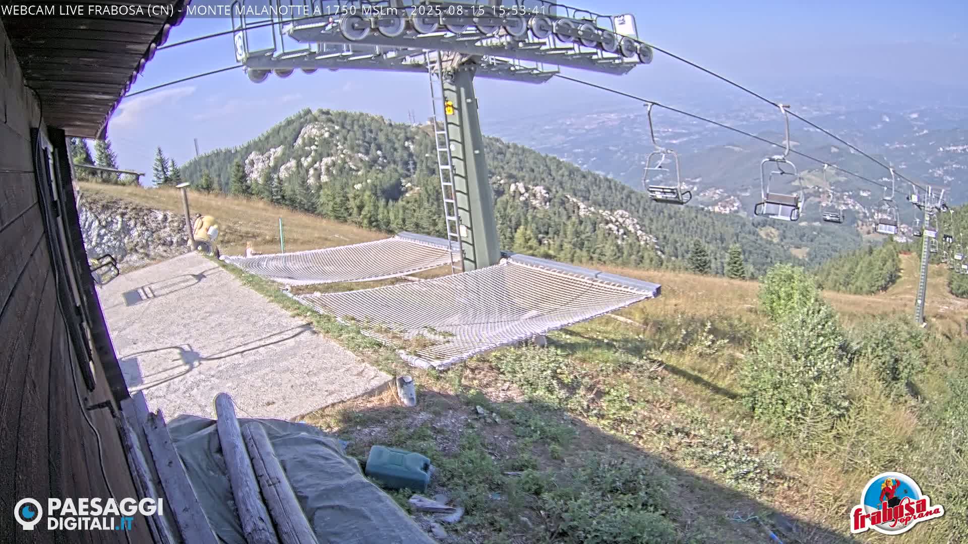 A mountaintop view on a sunny day shows a chairlift, two empty hammocks, and a gravel path next to a wooden building.