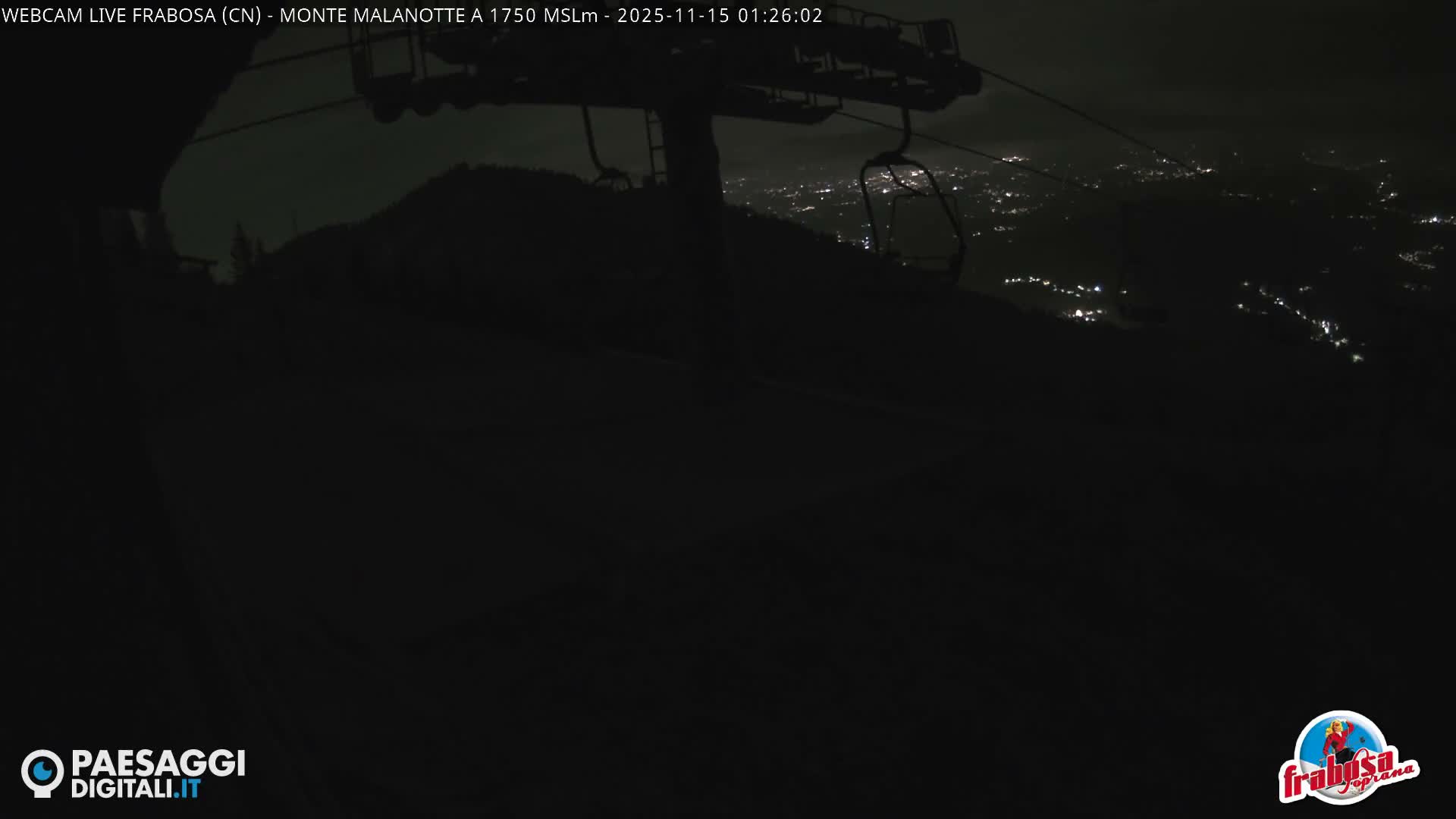 A dark night view from a mountain shows a ski lift in the foreground, with the distant, twinkling lights of a town spread across the valley below.