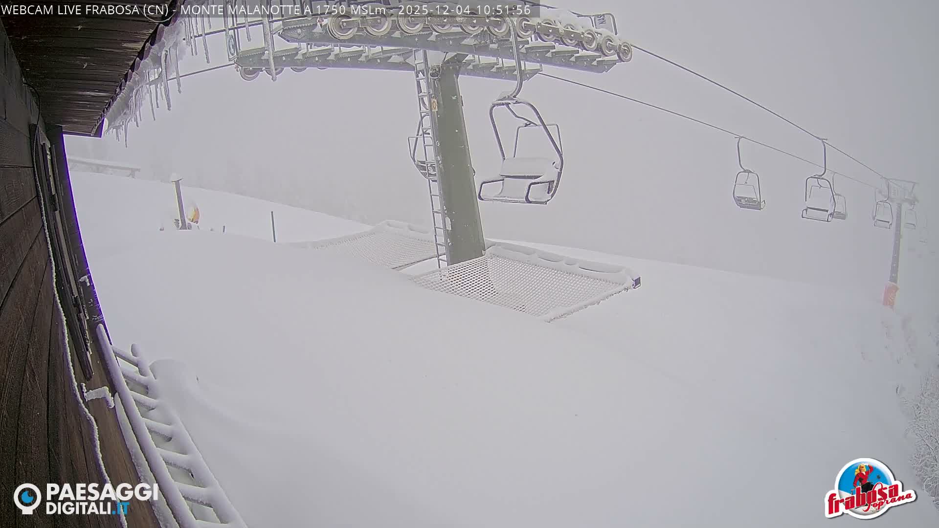 A snow-covered ski slope features an idle ski lift with empty chairs and a safety net near a wooden building adorned with icicles, all obscured by heavy snowfall and poor visibility.