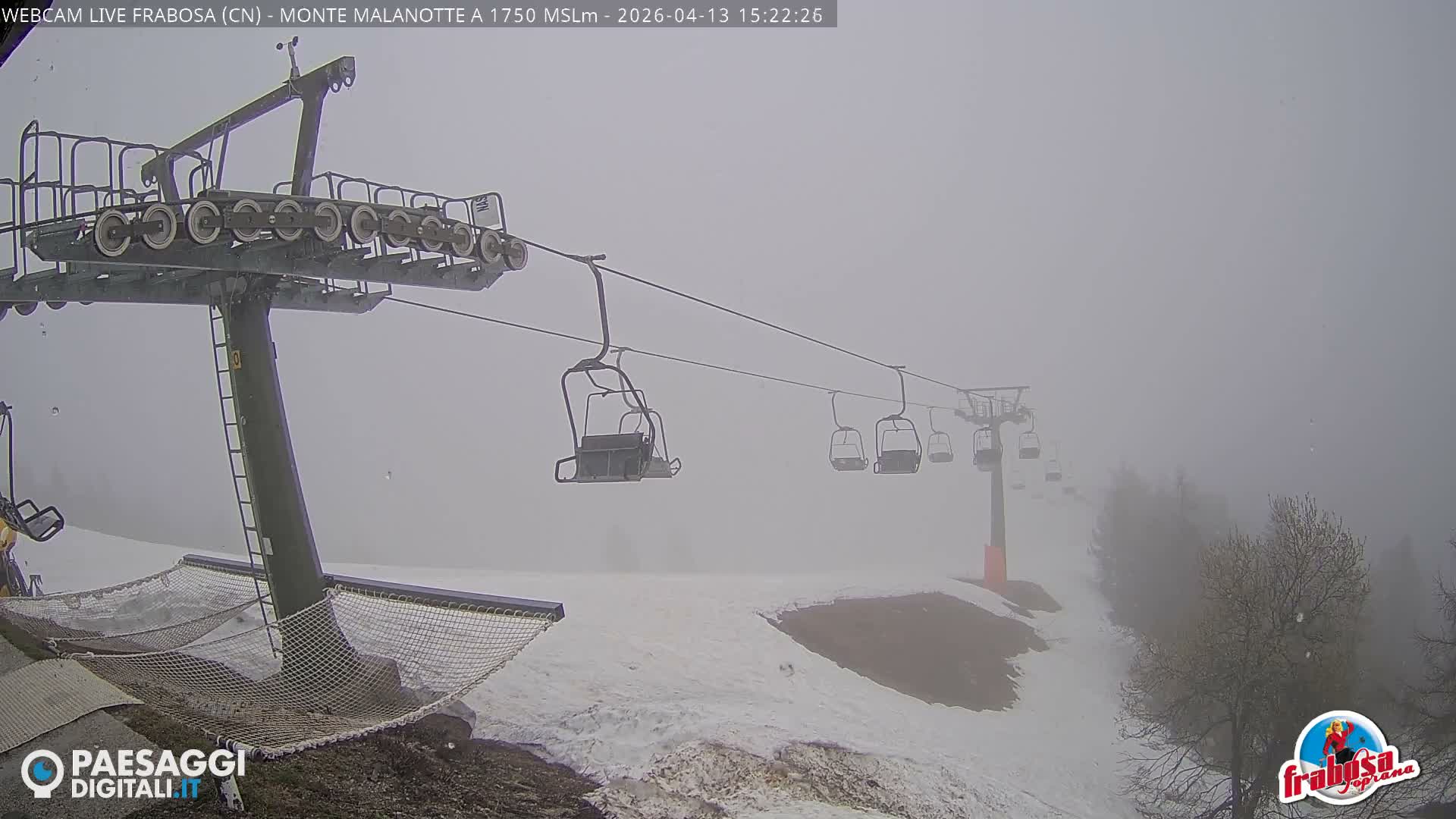 A snow-covered ski slope features an idle ski lift with empty chairs and a safety net near a wooden building adorned with icicles, all obscured by heavy snowfall and poor visibility.
