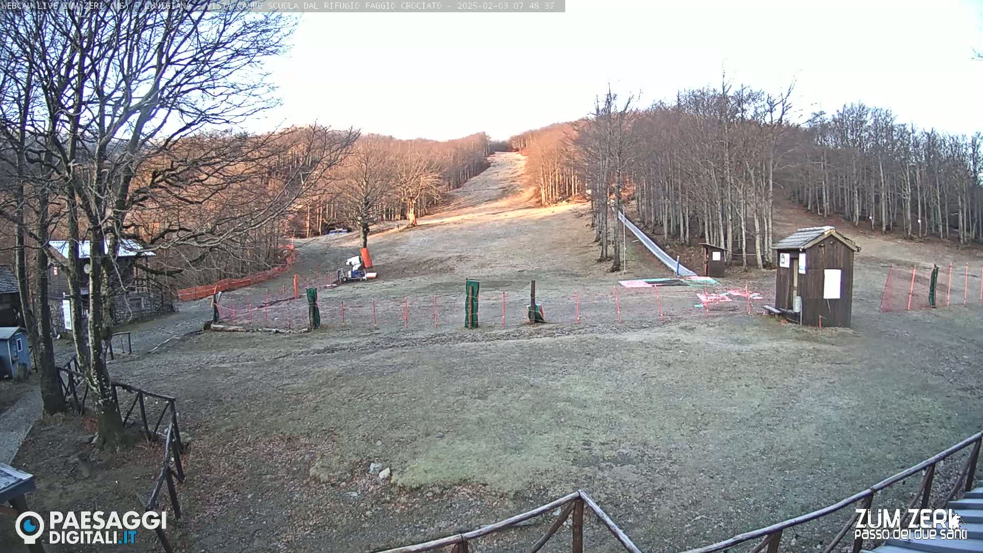 A mostly barren ski slope is visible, surrounded by leafless trees under a mostly clear sky.