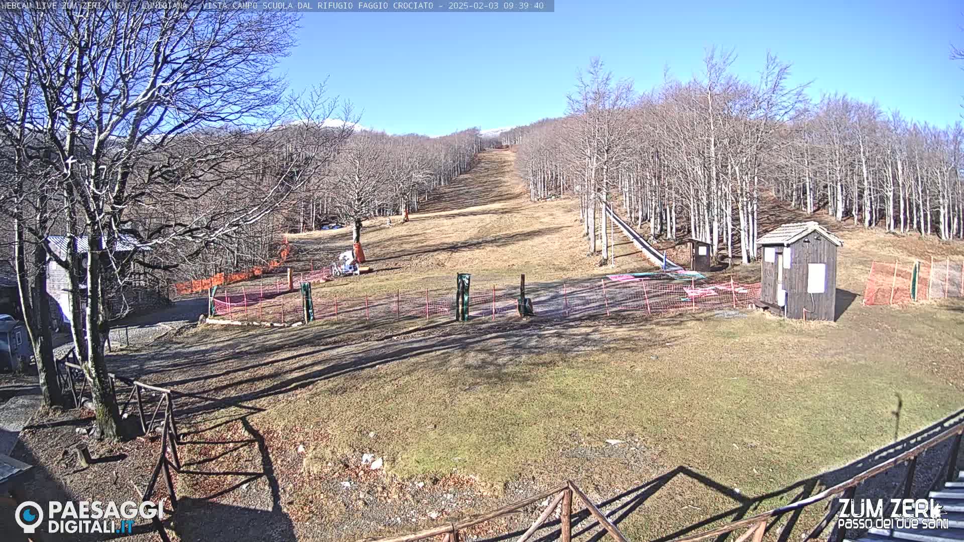 A sunny view of a ski slope with leafless trees, a small wooden building, and snow-covered mountains in the distance.