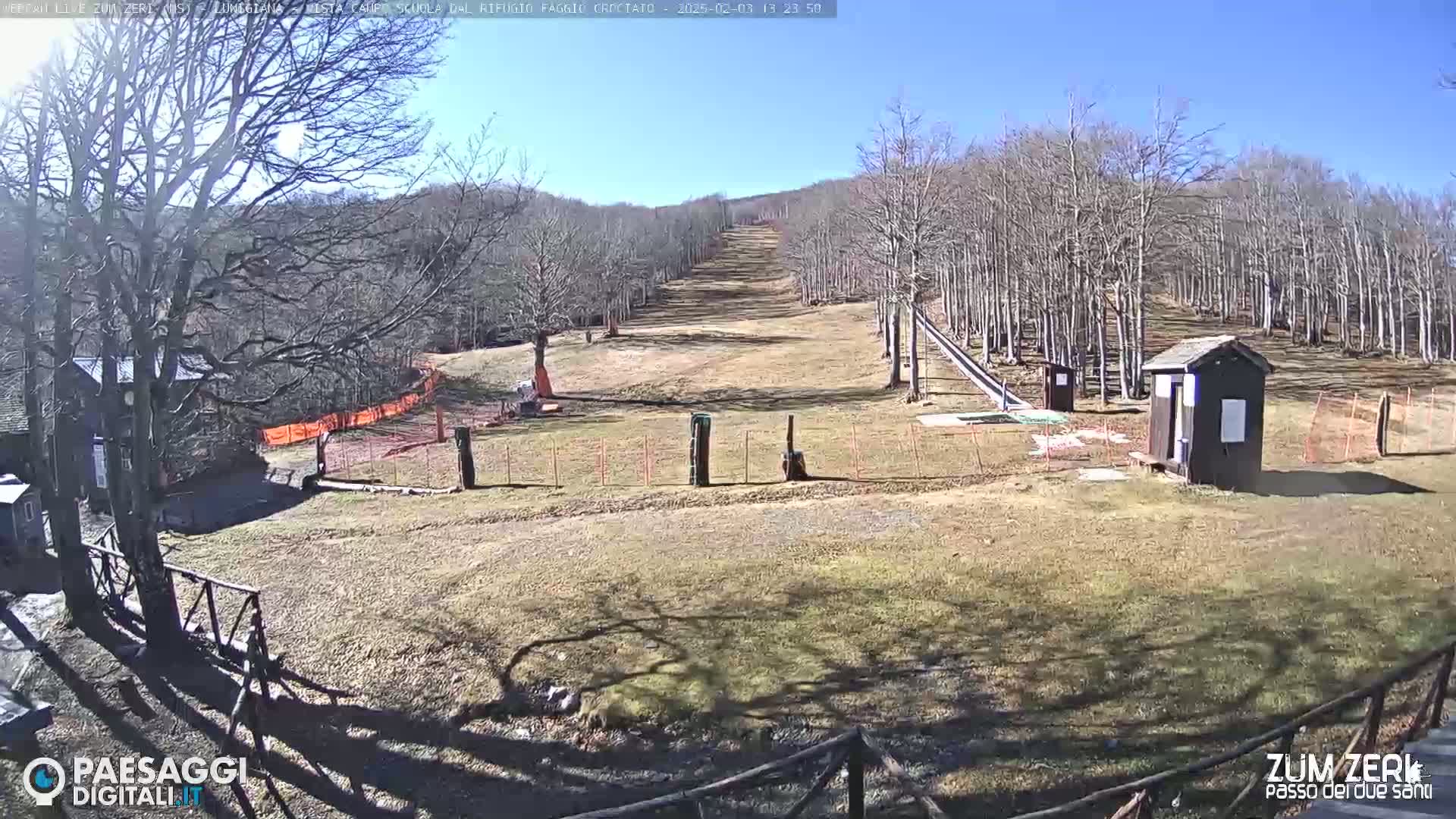 A sunny day at a ski slope shows a mostly bare, brown landscape with leafless trees and a small building.