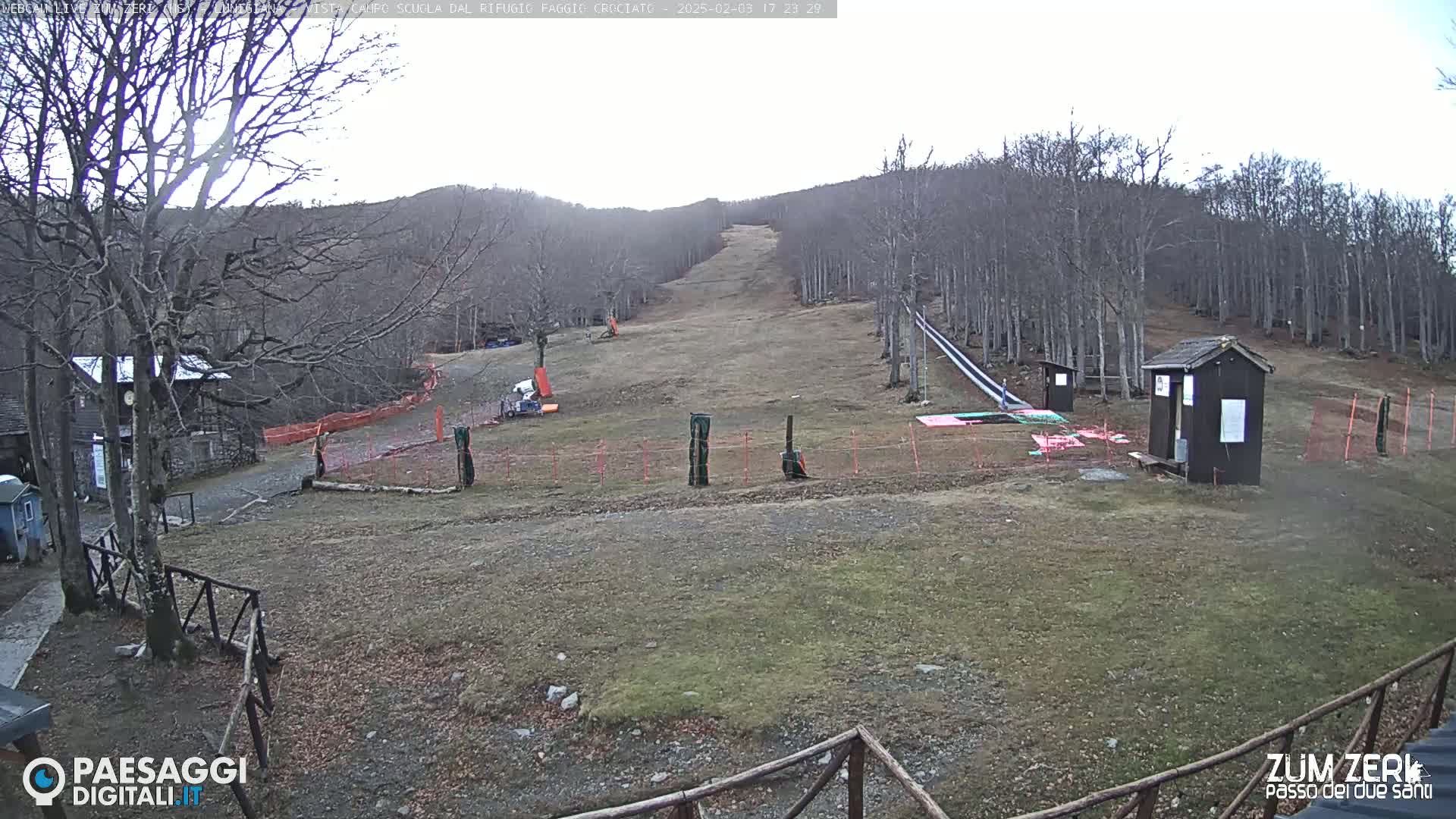 A mostly barren ski slope in a forest on an overcast day, with a few small buildings and equipment visible.