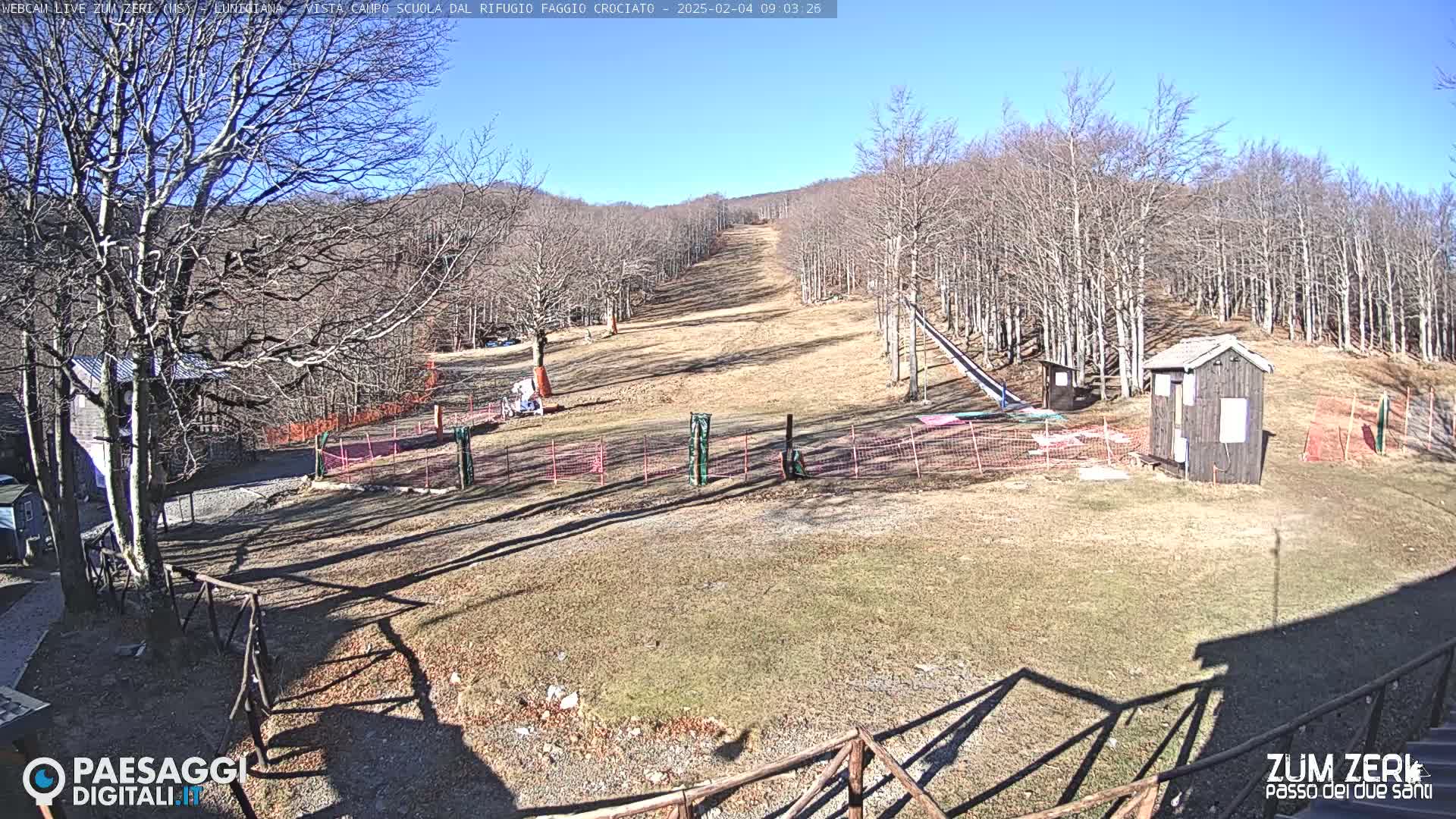 A sunny, snow-free ski slope is visible, surrounded by leafless trees and small structures.