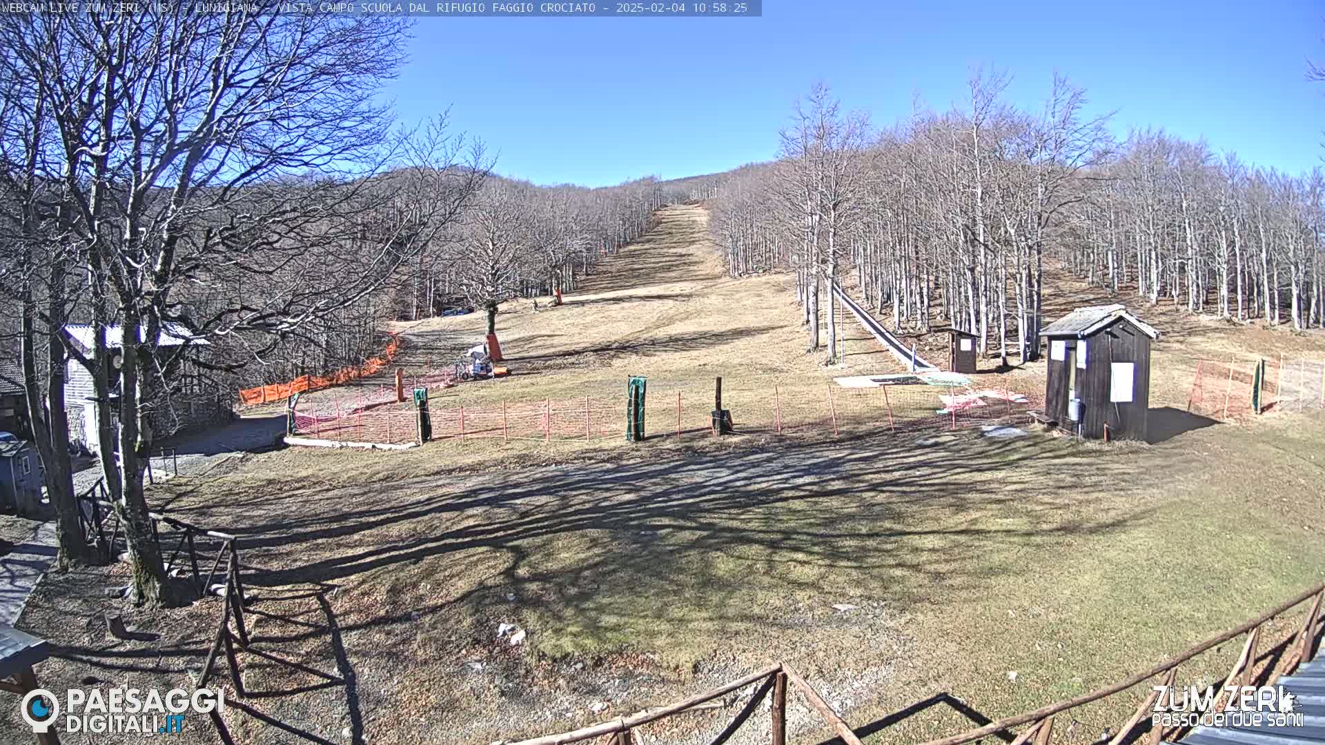 A mostly sunny view of a ski slope in a wooded area, with several small buildings and orange fencing.