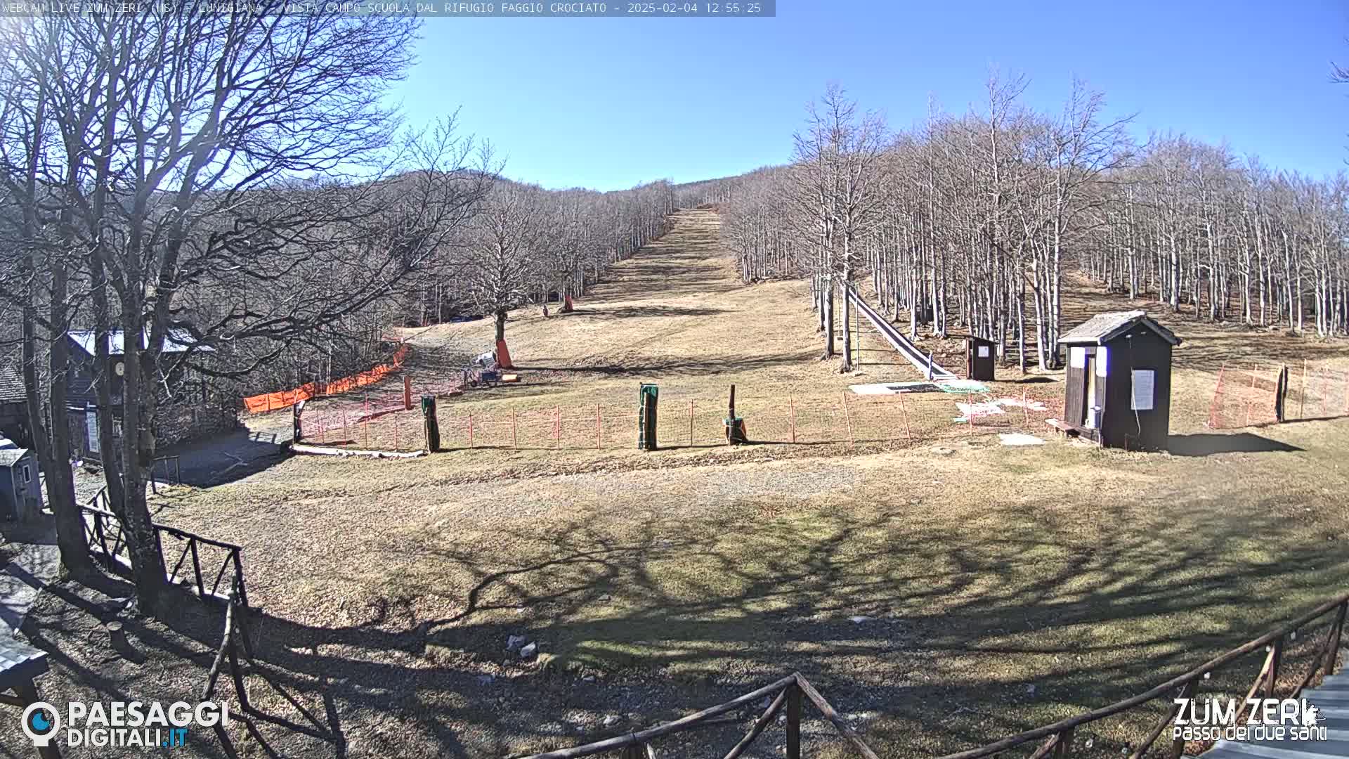 A mostly sunny day reveals a sparsely populated ski slope with leafless trees, small buildings, and orange safety netting.