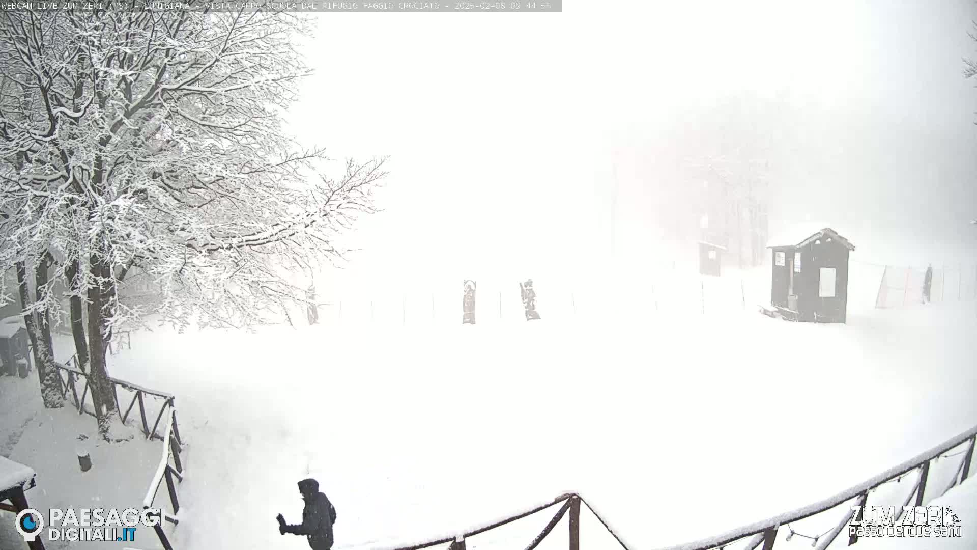A snow-covered landscape with a person walking near a wooden fence and a small structure in the distance, during a heavy snowfall.
